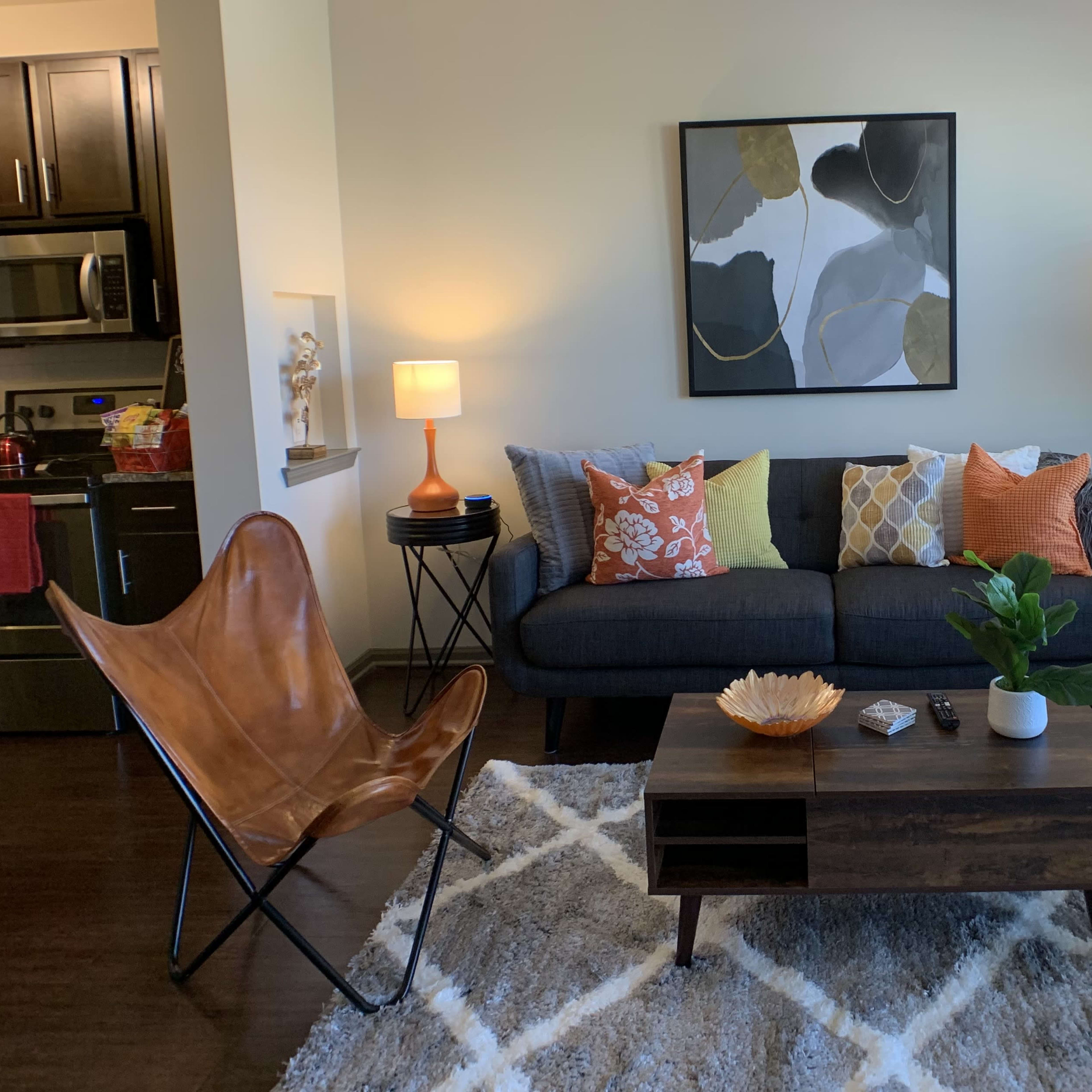 A living room with a dark sofa adorned with colorful pillows, a wooden coffee table, a decorative bowl, a plant, and a soft area rug, all set against light-colored walls and a window with blinds.