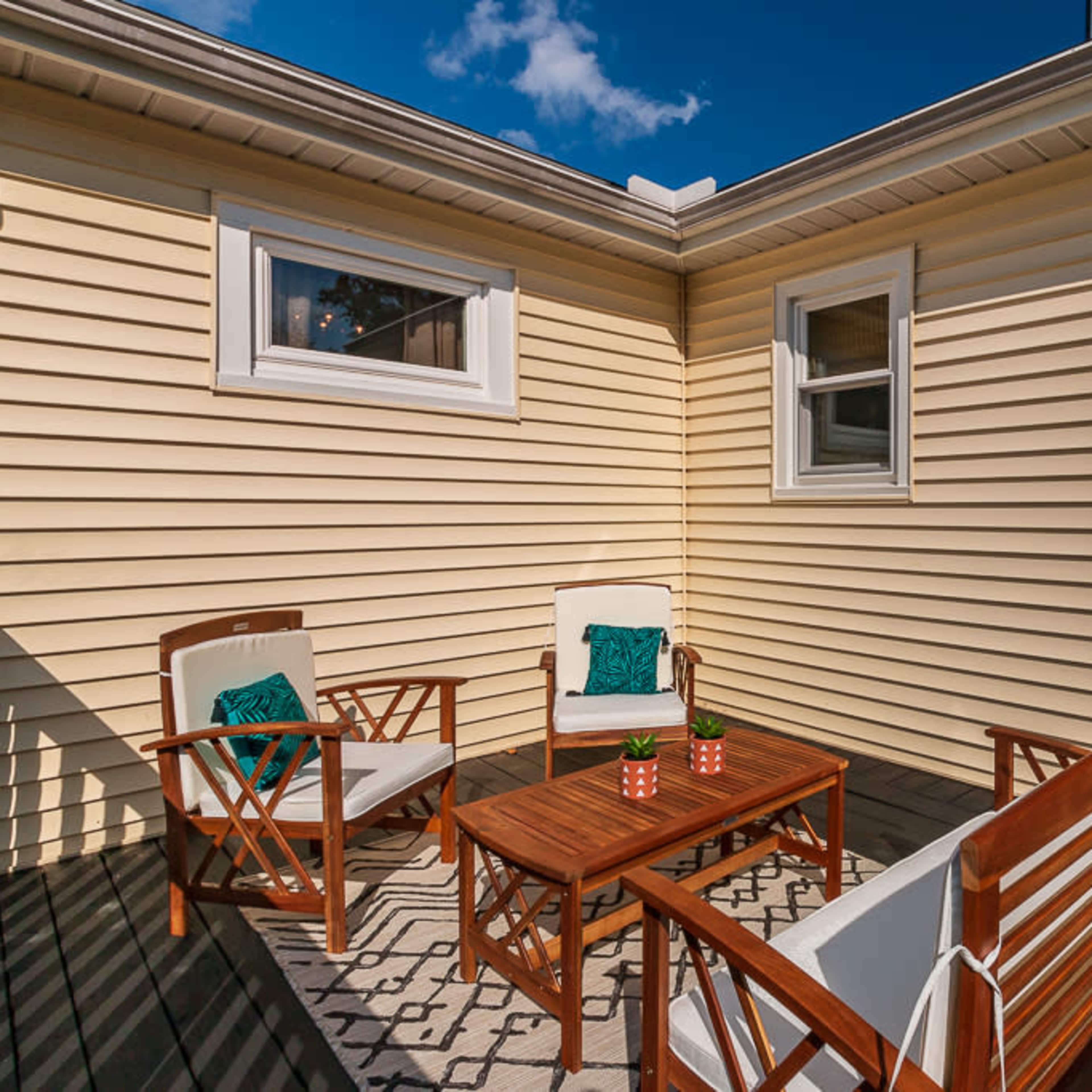A small outdoor patio features two wooden chairs and a table, surrounded by yellow siding and a single door.