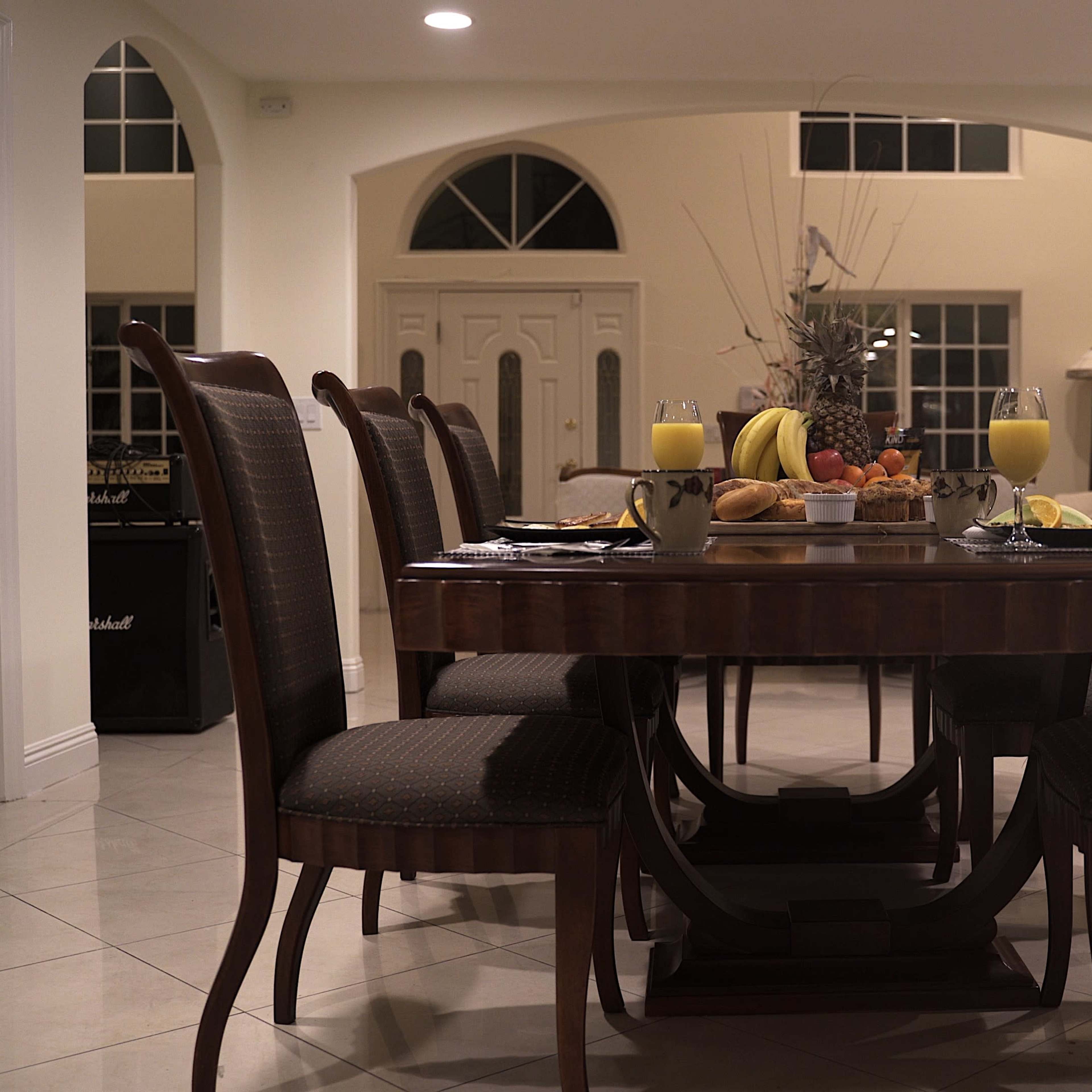 A dining table is set with fruits and beverages, surrounded by chairs in a well-lit room with large windows.