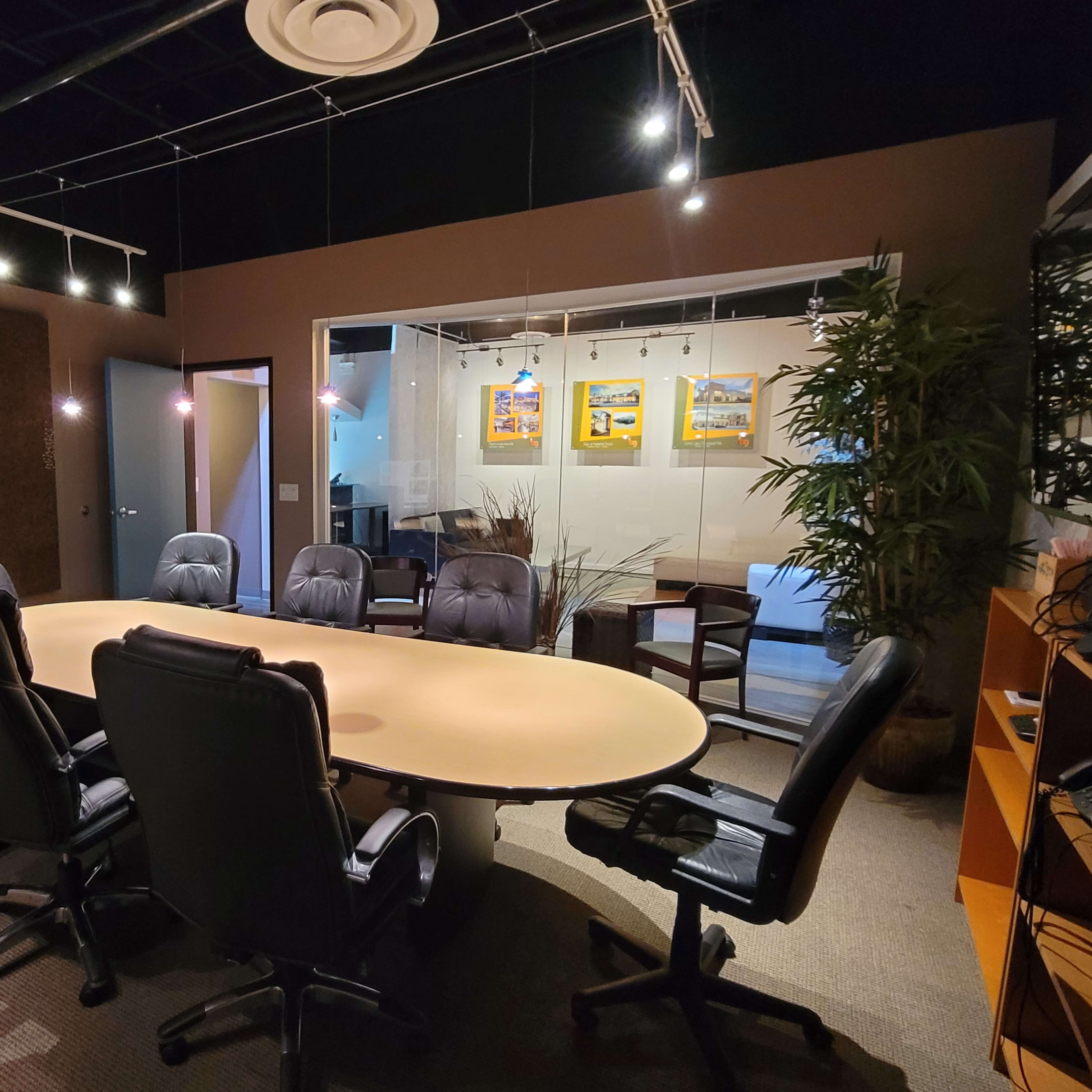 A conference room features a large oval table surrounded by black office chairs, with a glass wall showcasing artwork and a seating area in the background.