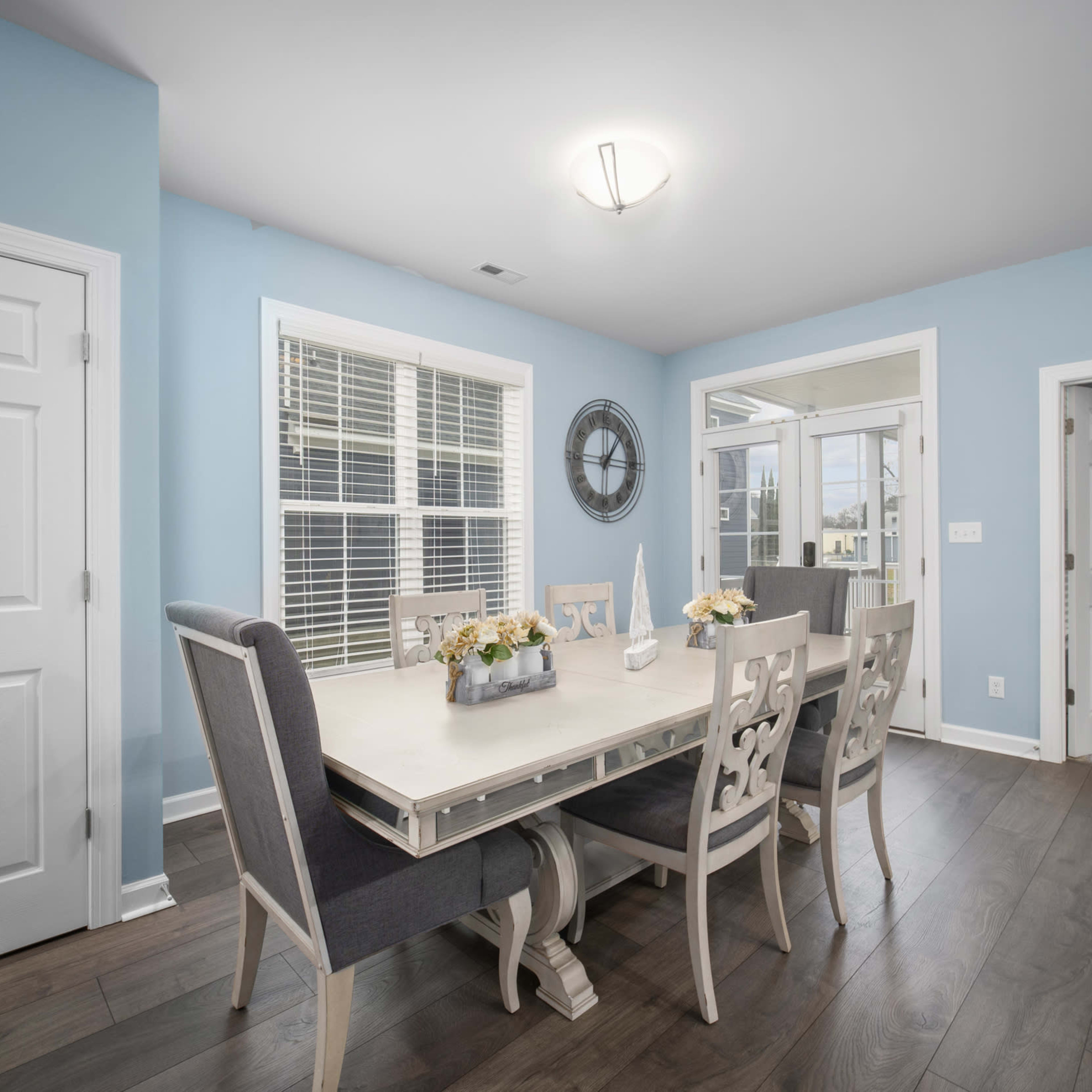 A dining area with a light blue wall, a large table surrounded by upholstered chairs, and doors leading to an outdoor space.