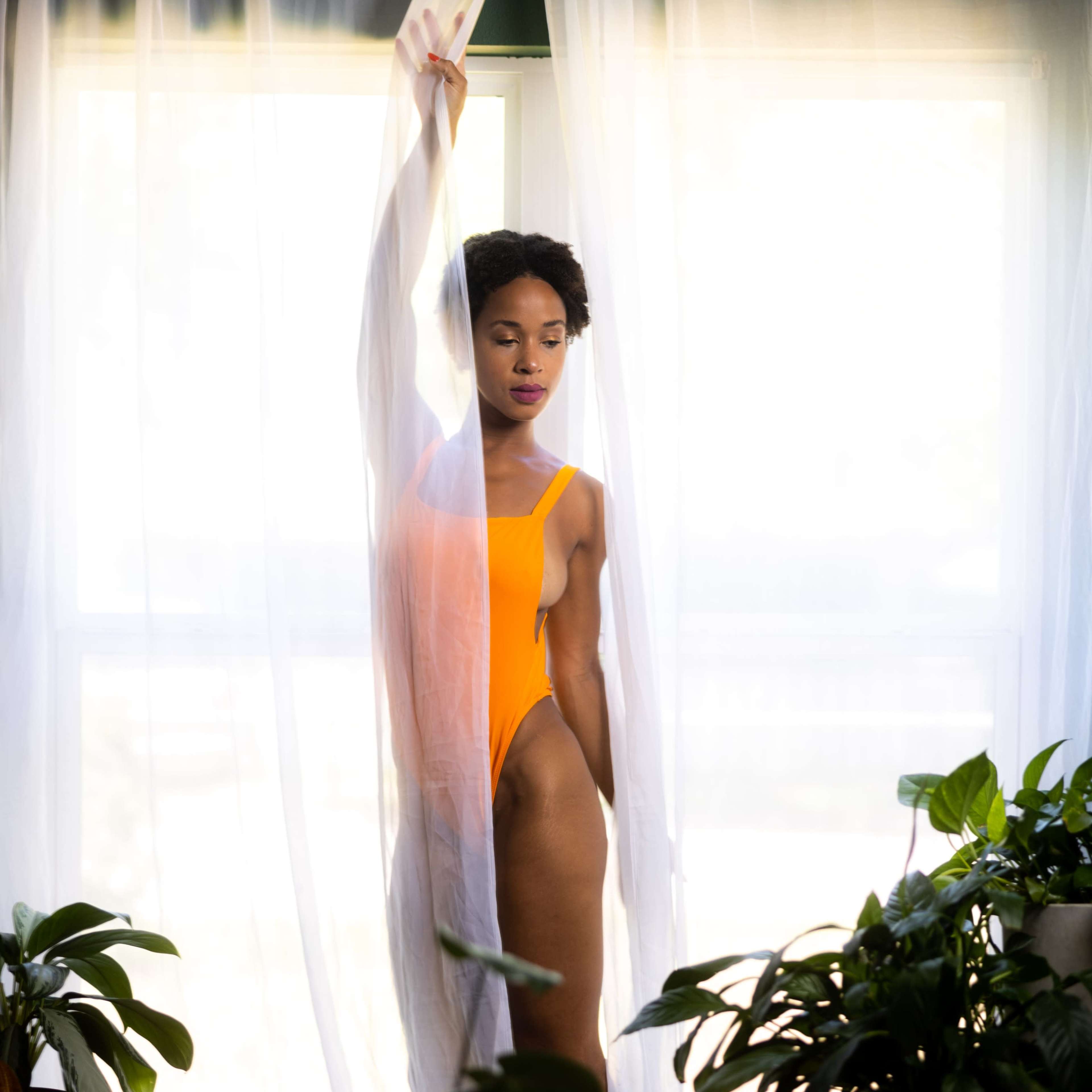 A person in an orange swimsuit stands by a window, partially obscured by a sheer curtain and surrounded by potted plants.