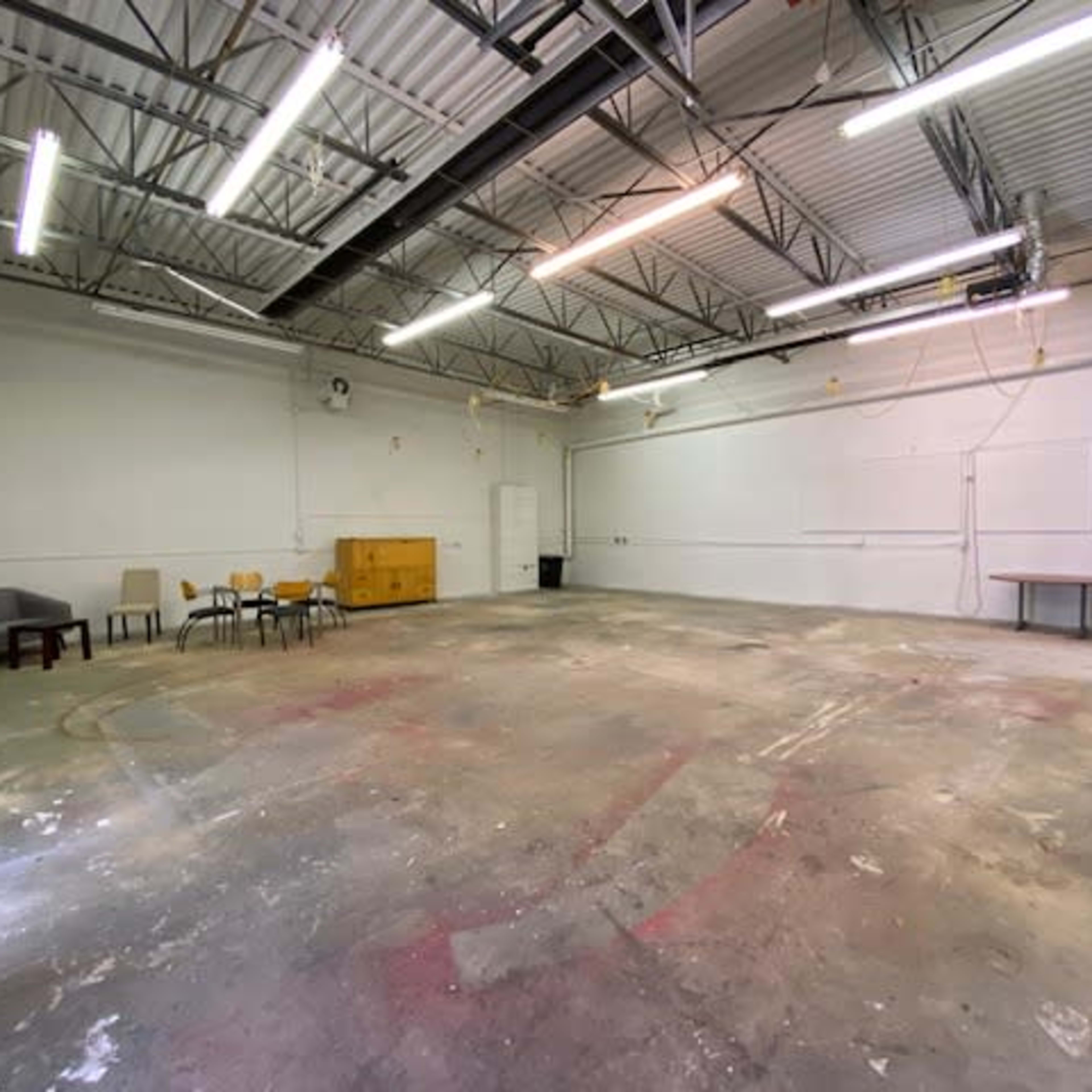An empty industrial space features a concrete floor, scattered furniture, and overhead lighting.
