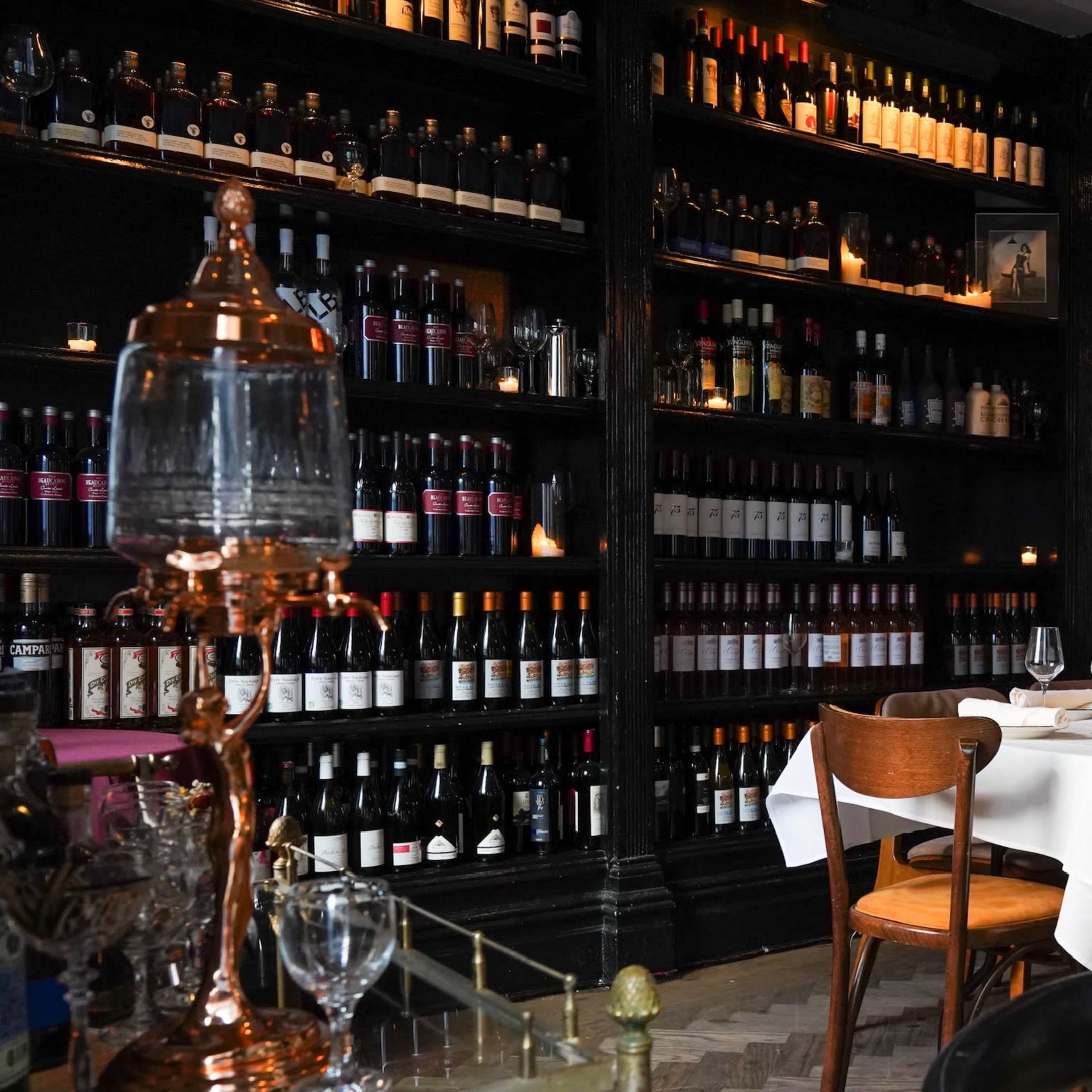A neatly set dining table is positioned in front of a tall black bookshelf filled with various bottles of wine in a restaurant.