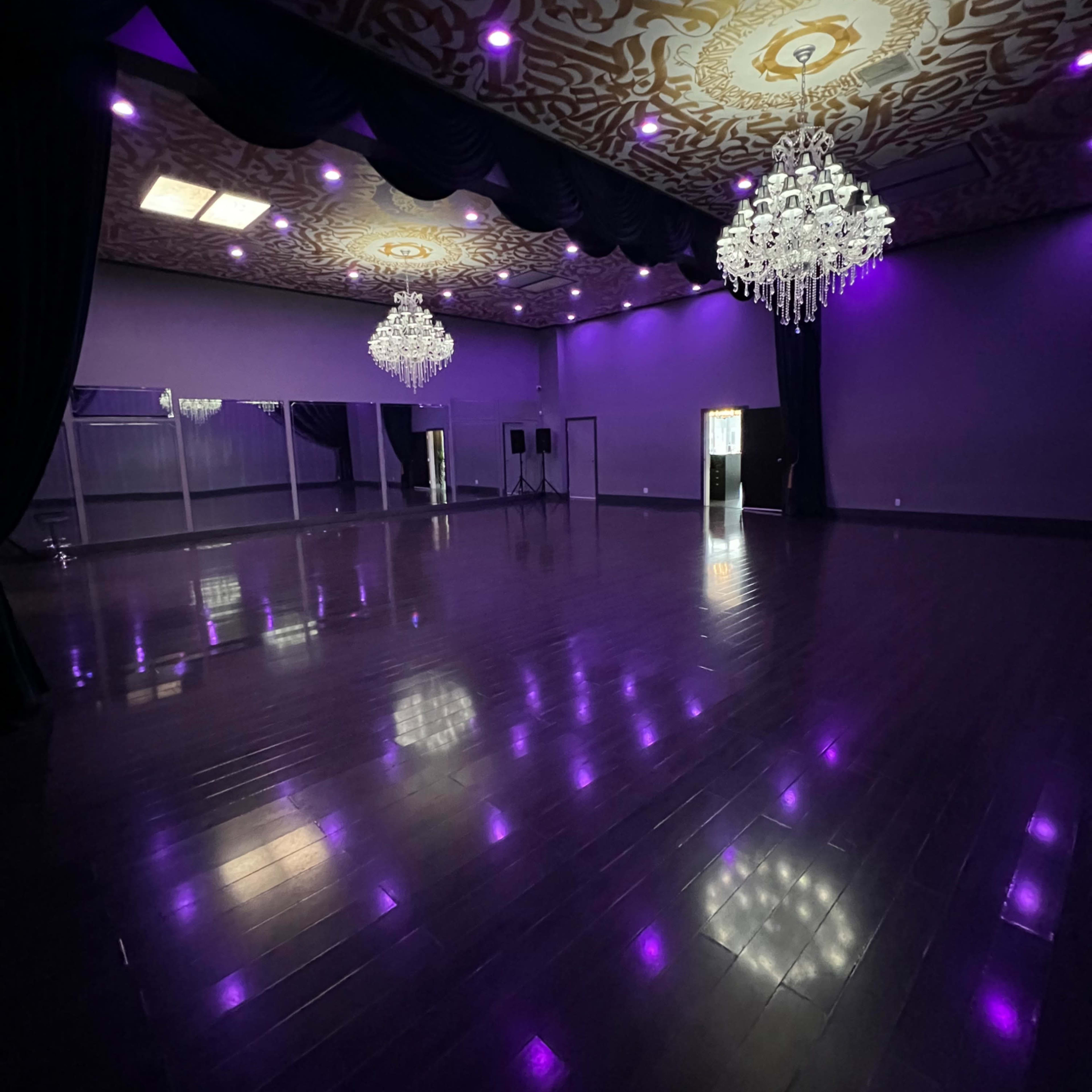 A large, empty ballroom with polished wooden floors, purple walls, and crystal chandeliers hanging from the ceiling.
