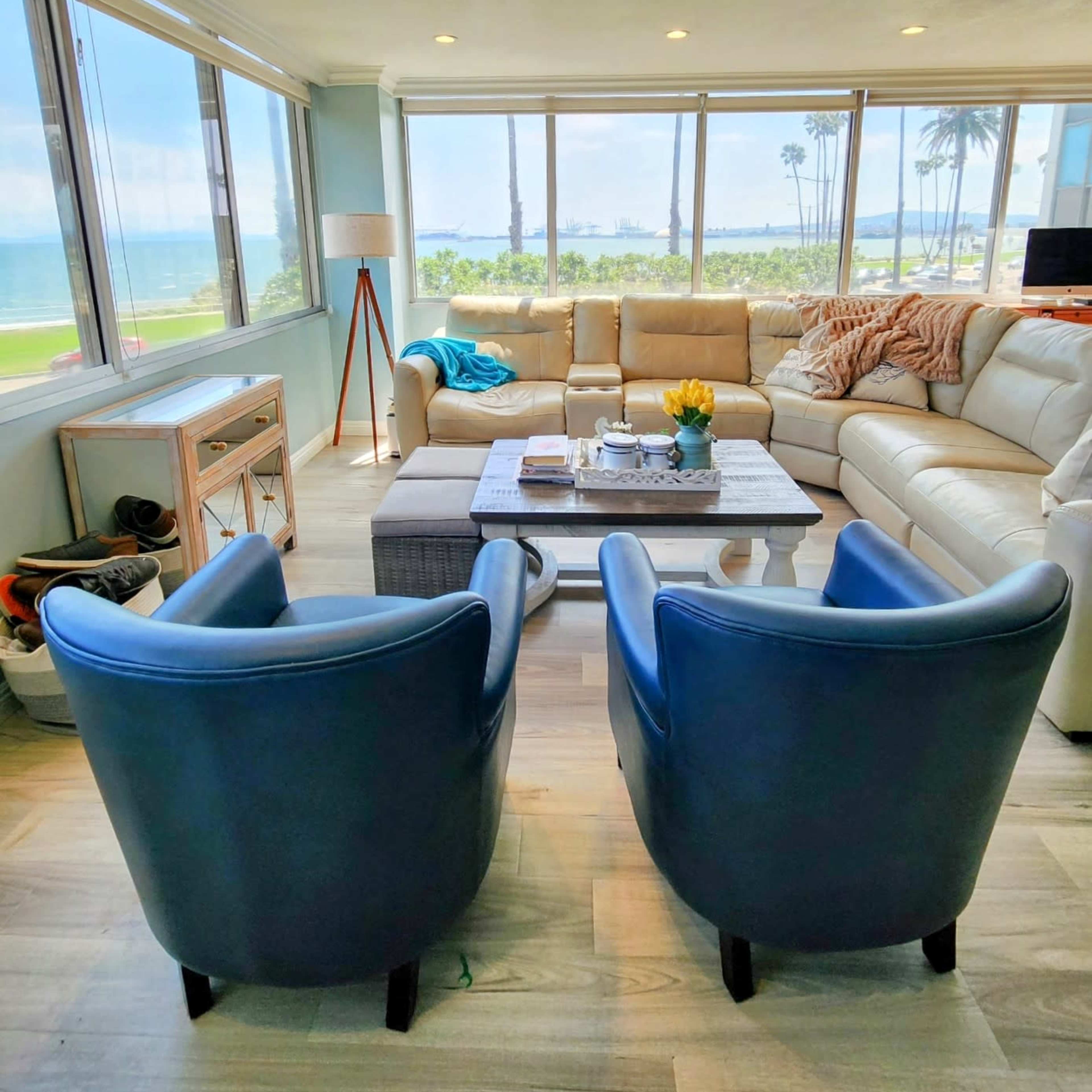 The scene shows a bright living room with a large sectional sofa and two blue chairs, overlooking a beach and palm trees through large windows.