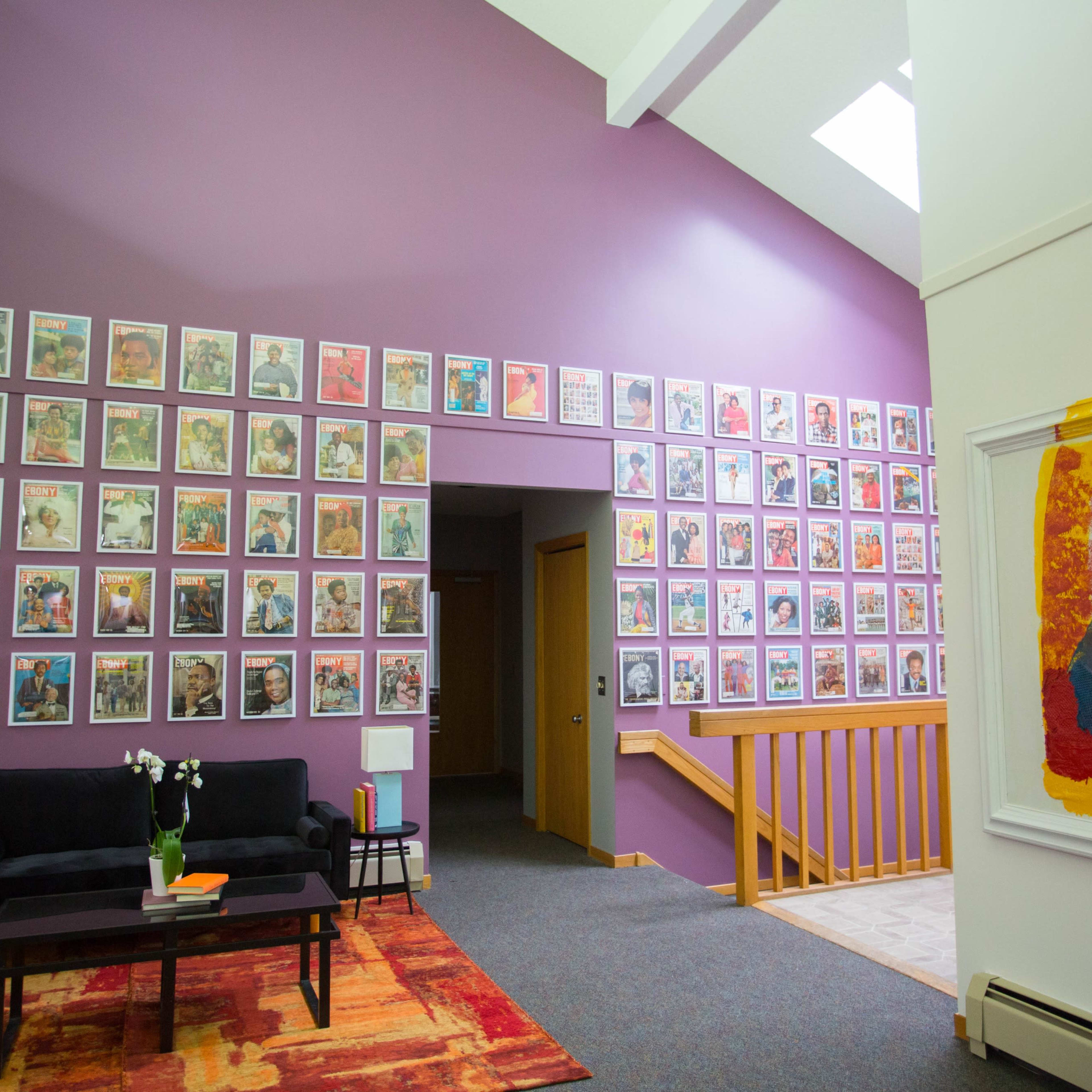 A brightly lit interior space featuring a wall adorned with numerous framed artworks in vibrant colors, complemented by a black sofa and wooden staircase.