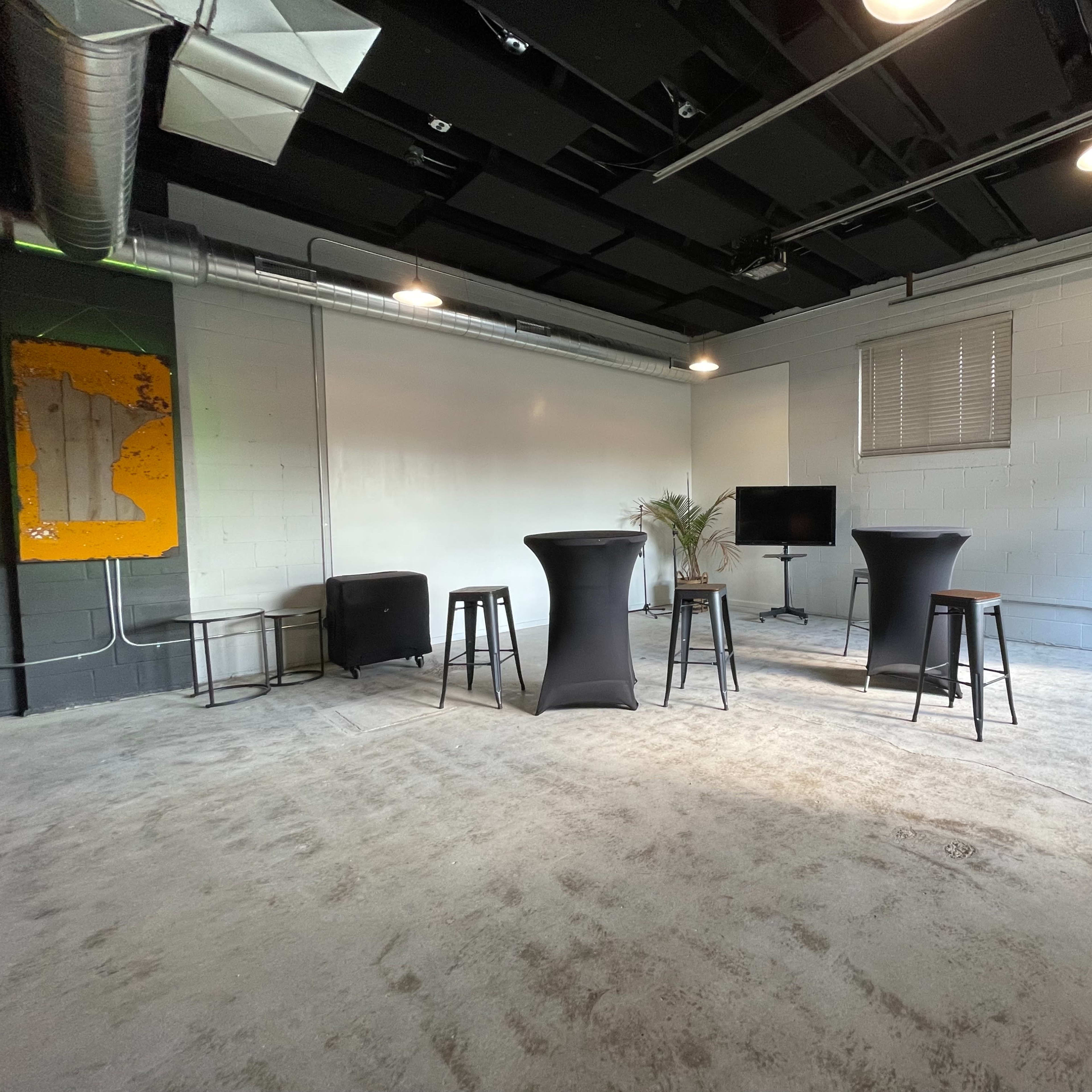 A sparse room with a concrete floor, featuring two tall black tables, a television on a stand, and a leafy plant near the back wall.