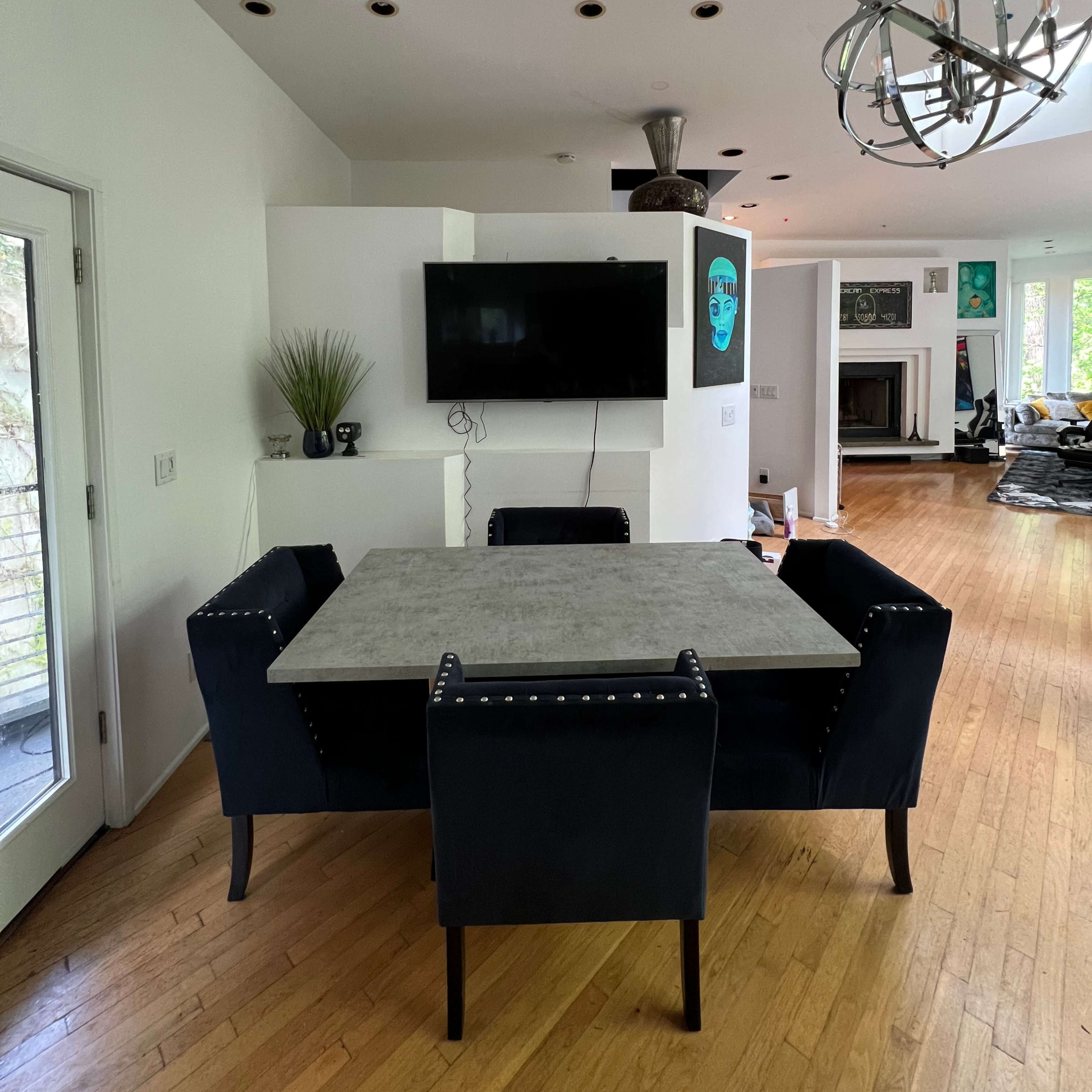 A dining area features a rectangular table surrounded by four upholstered chairs, with a wall-mounted television and an overhead light fixture above.
