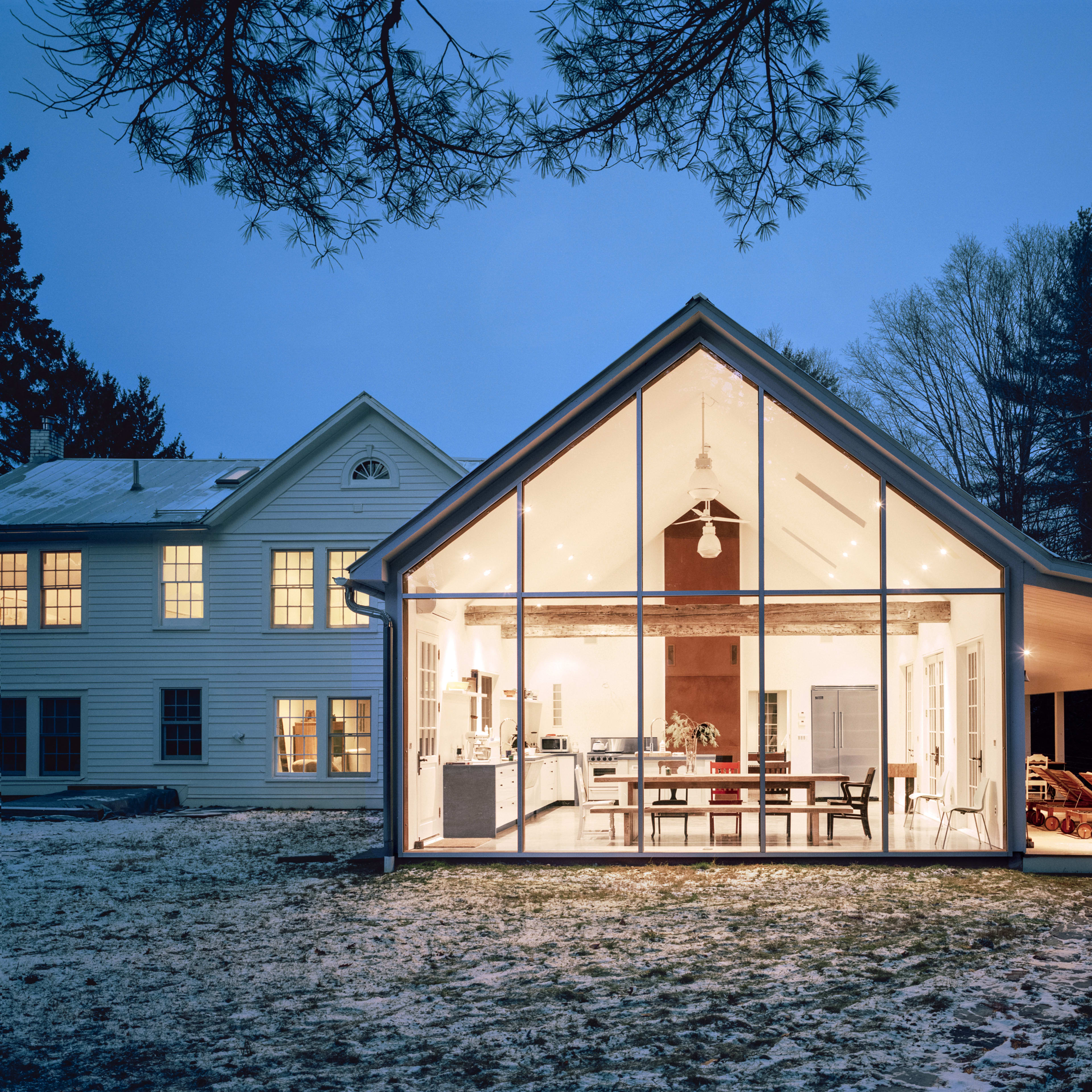 A modern, glass-fronted extension of a house is illuminated at dusk, showcasing a spacious interior with a dining area and kitchen.