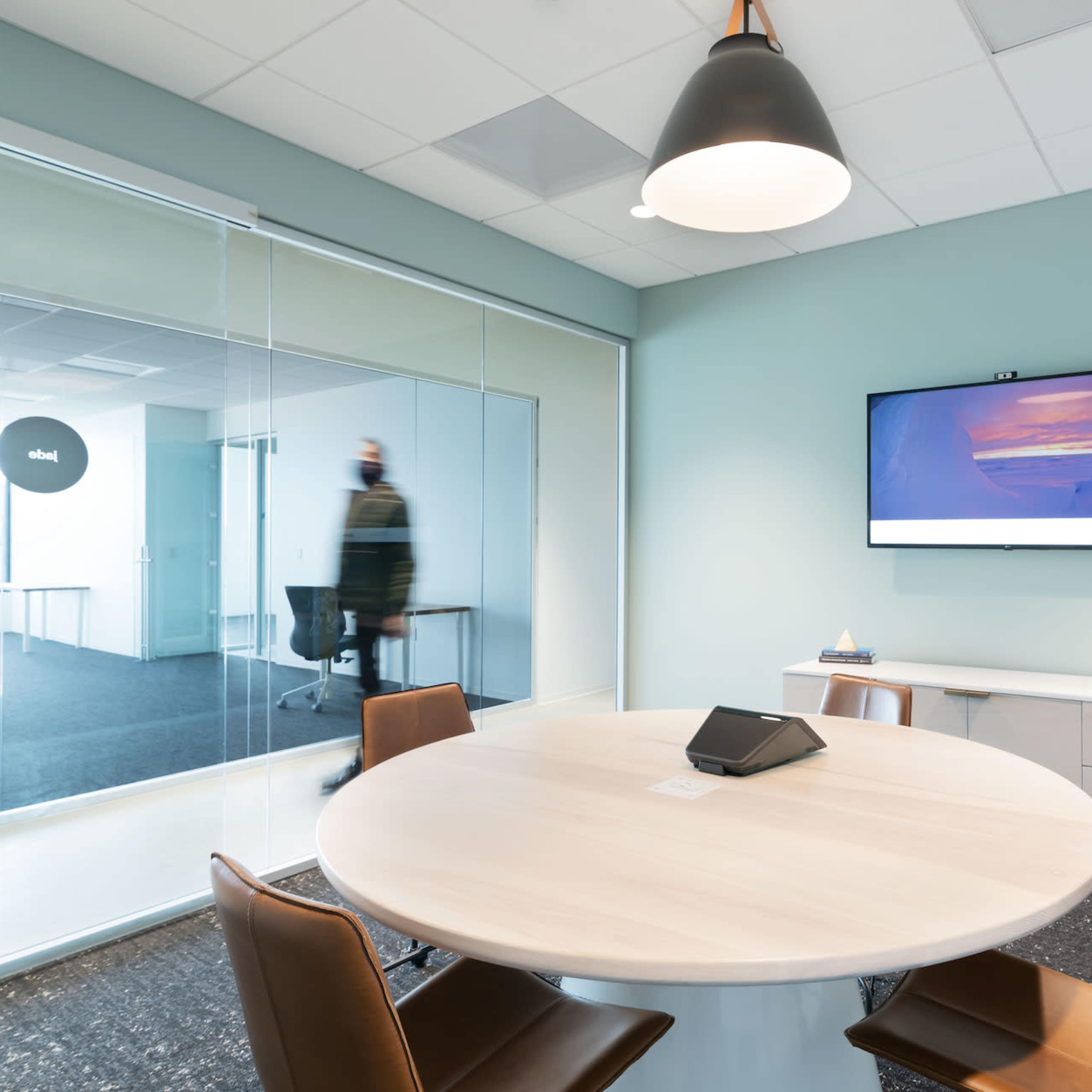 A modern conference room features a round table with leather chairs, a wall-mounted screen displaying an image, and a glass wall separating it from an adjacent space.
