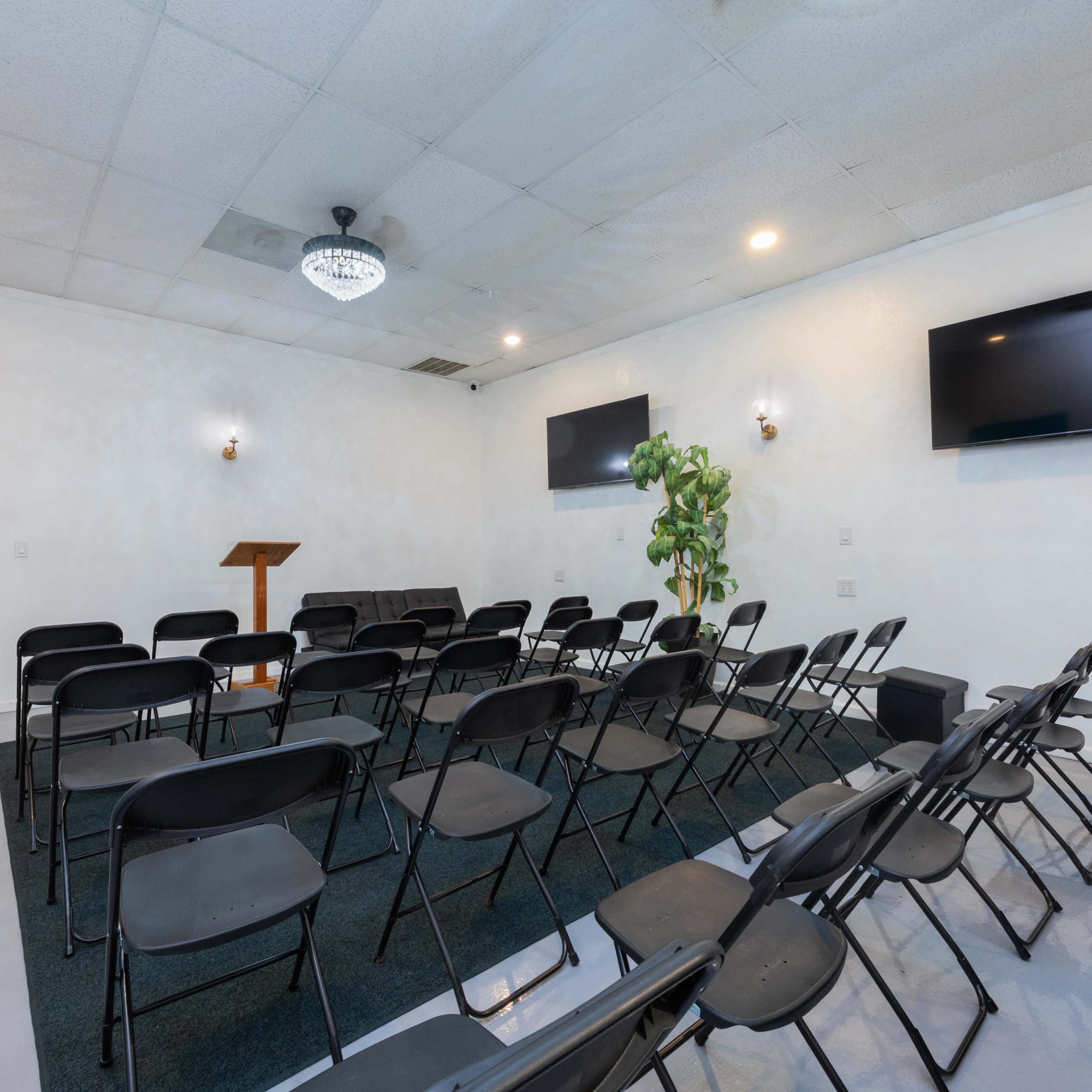 A small, empty room set up with rows of black chairs facing a wooden podium, flanked by two wall-mounted monitors and a potted plant in one corner.
