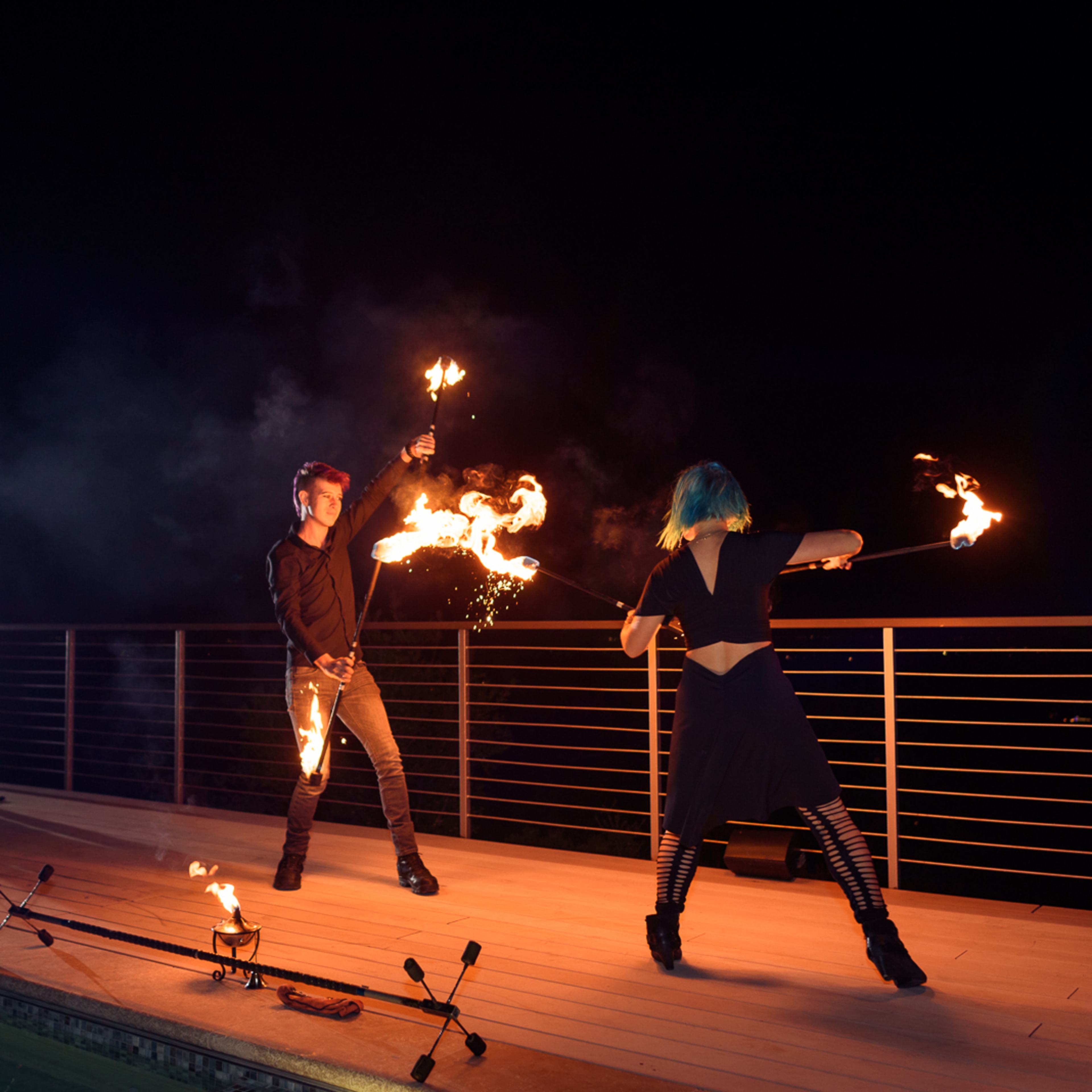 Two performers with flaming staffs dance on a wooden deck illuminated by lights at night, while a seated person watches in the background.