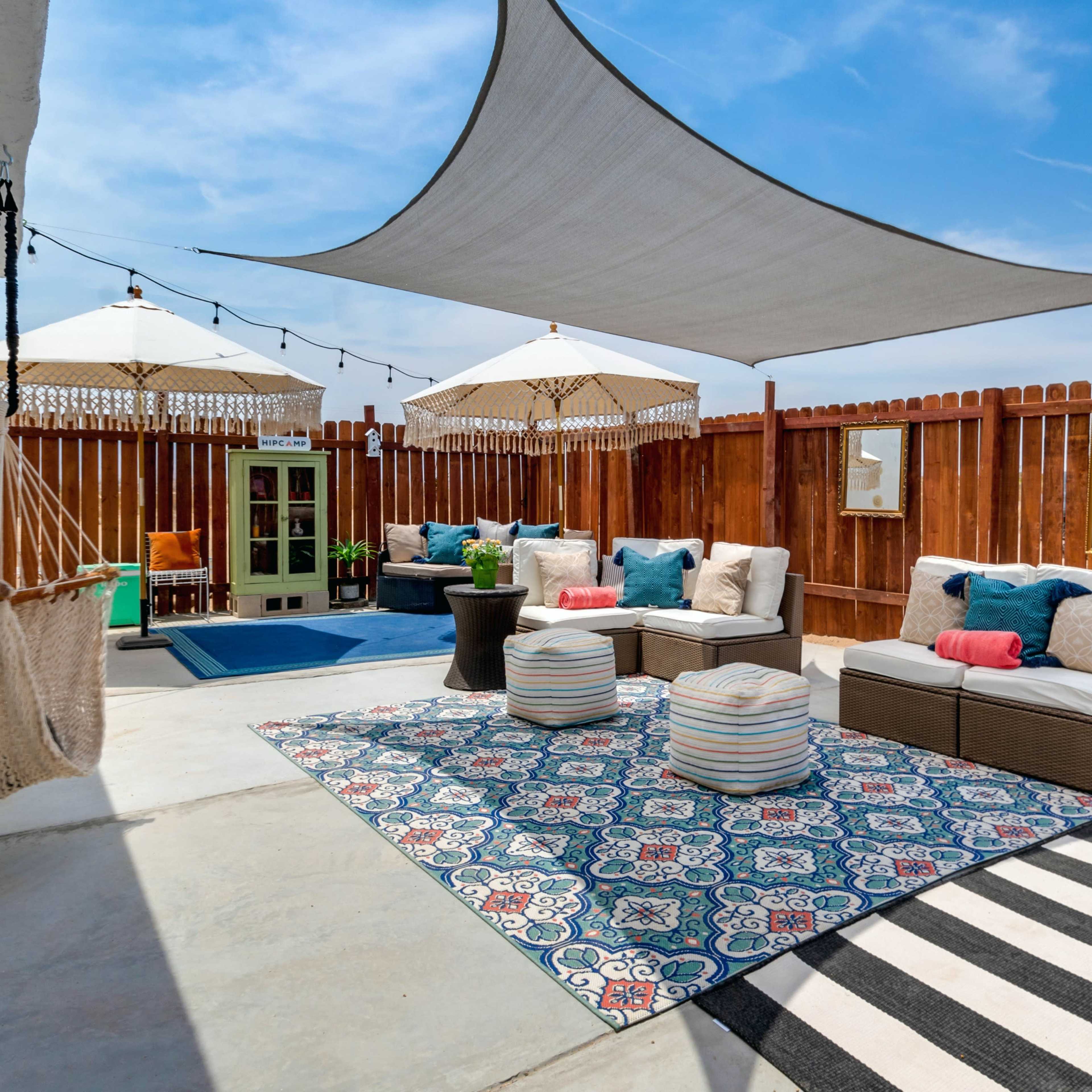 A patio area features a shaded seating arrangement with plush cushions, decorative umbrellas, and a patterned rug, surrounded by a wooden fence.