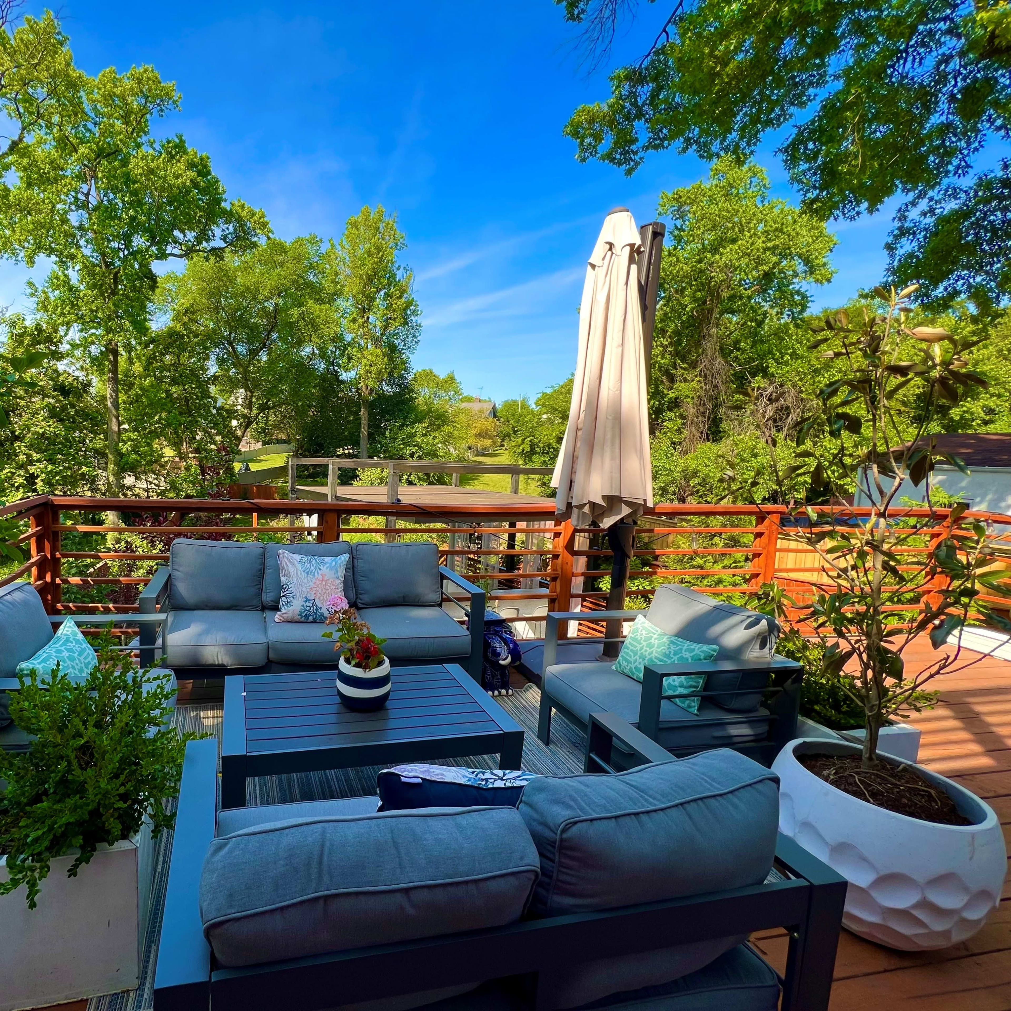 A comfortable outdoor seating area with couches and a large umbrella is surrounded by greenery and a wooden deck.