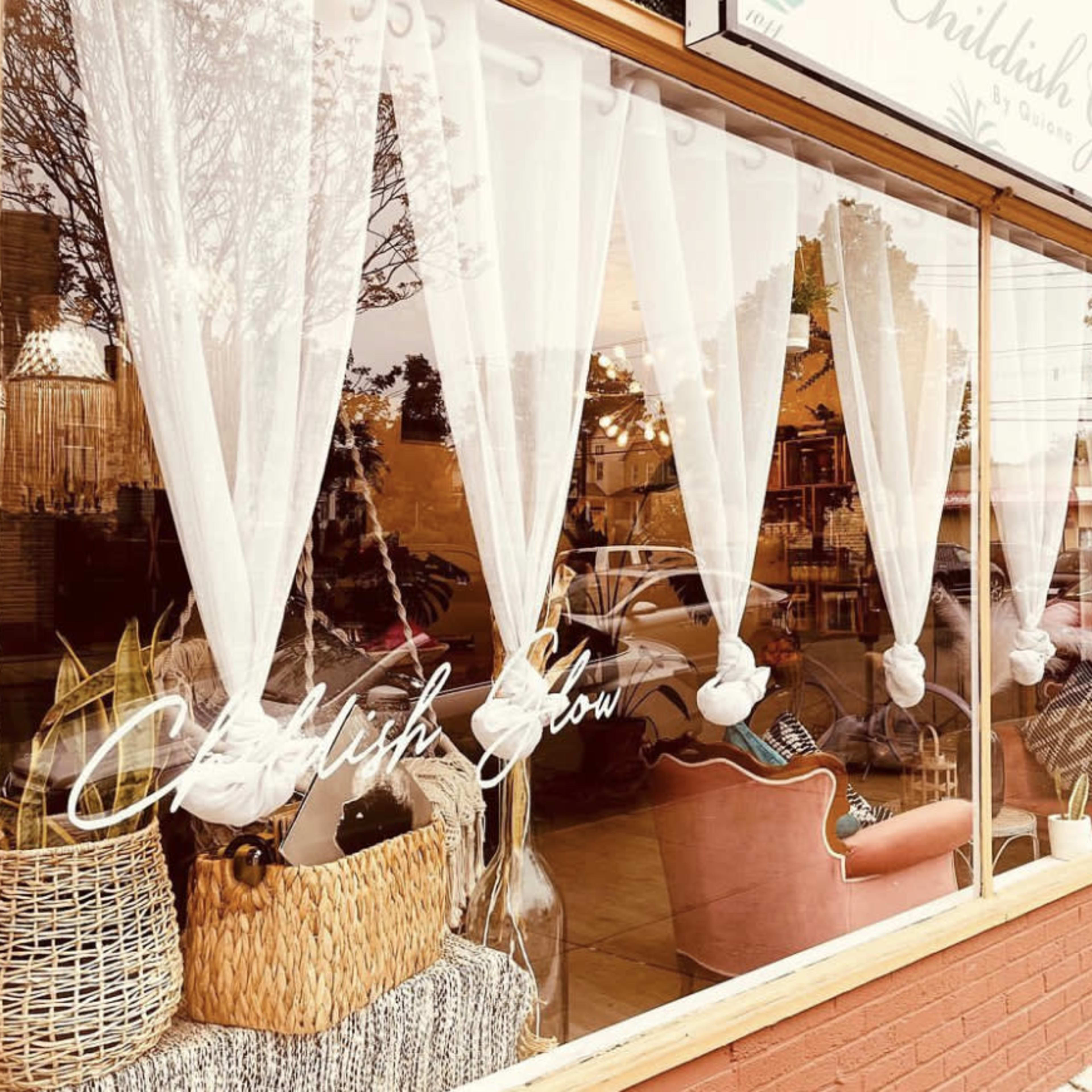 A storefront with large windows featuring white curtains, decorative wicker baskets, and a cozy pink chair inside, along with the name "Childish Glow" displayed on the glass.