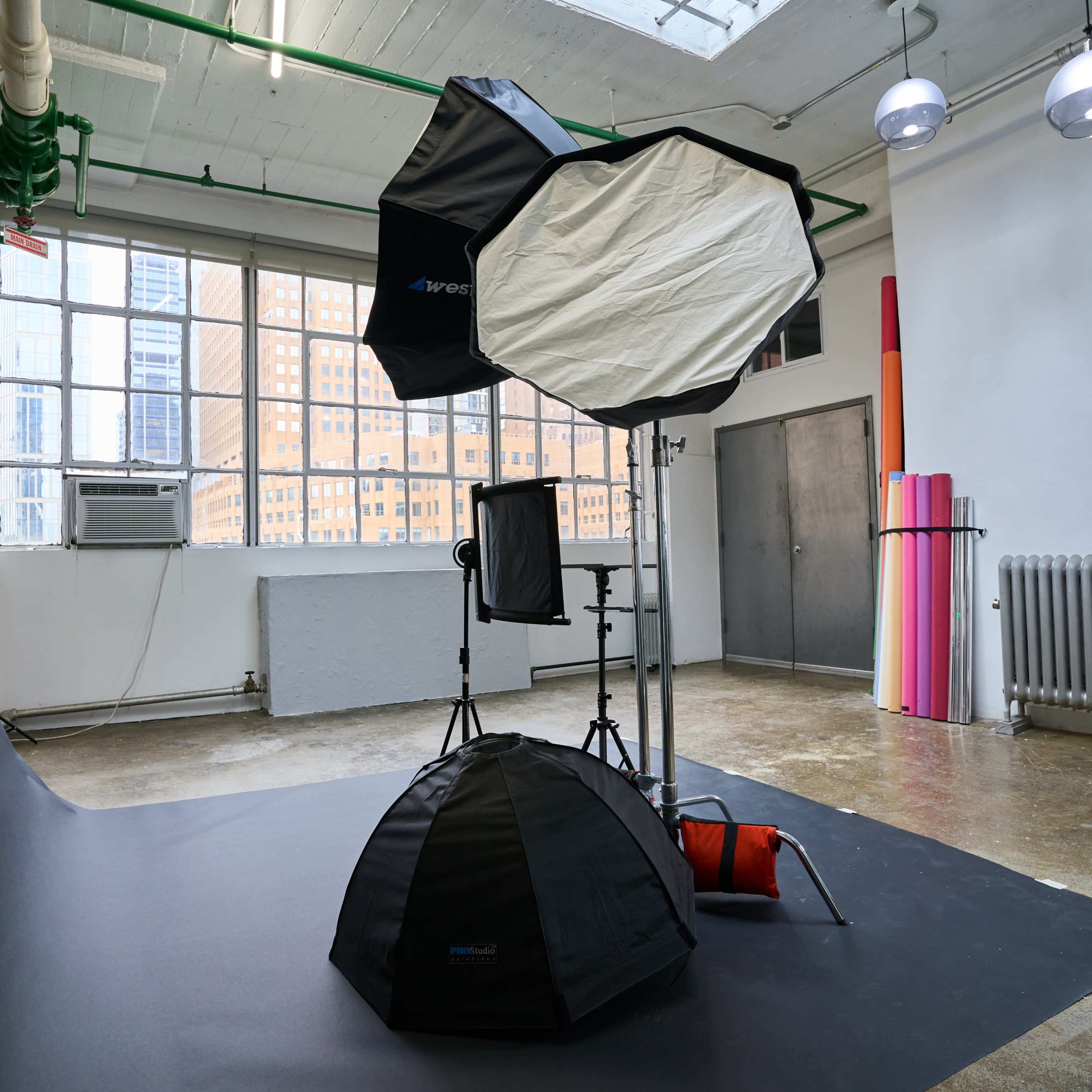 A photography studio setup with softbox lights, a backdrop, and various equipment against a white wall and large windows.