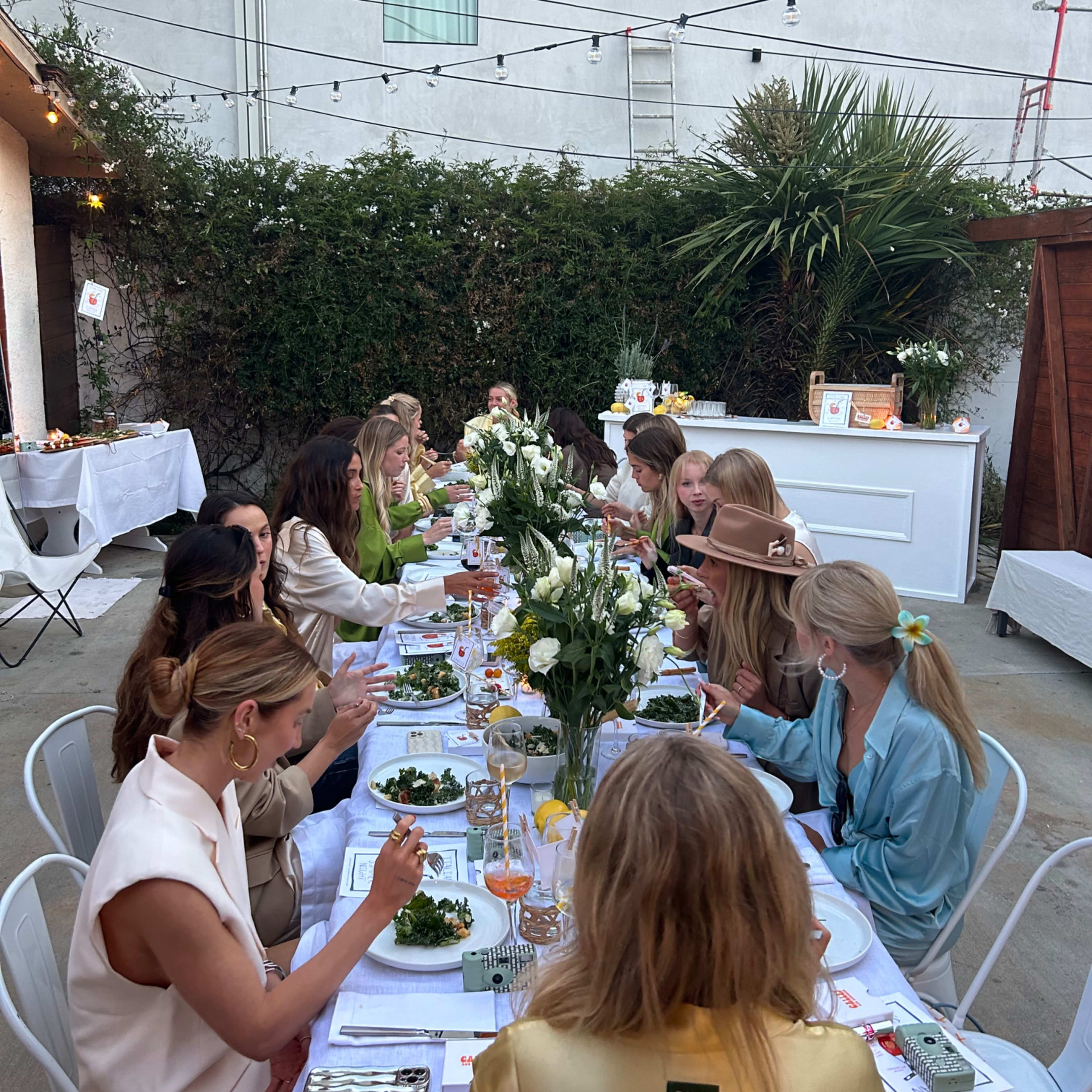 A long dining table is set outdoors with women dressed in various outfits, enjoying a meal amid greenery and string lights.
