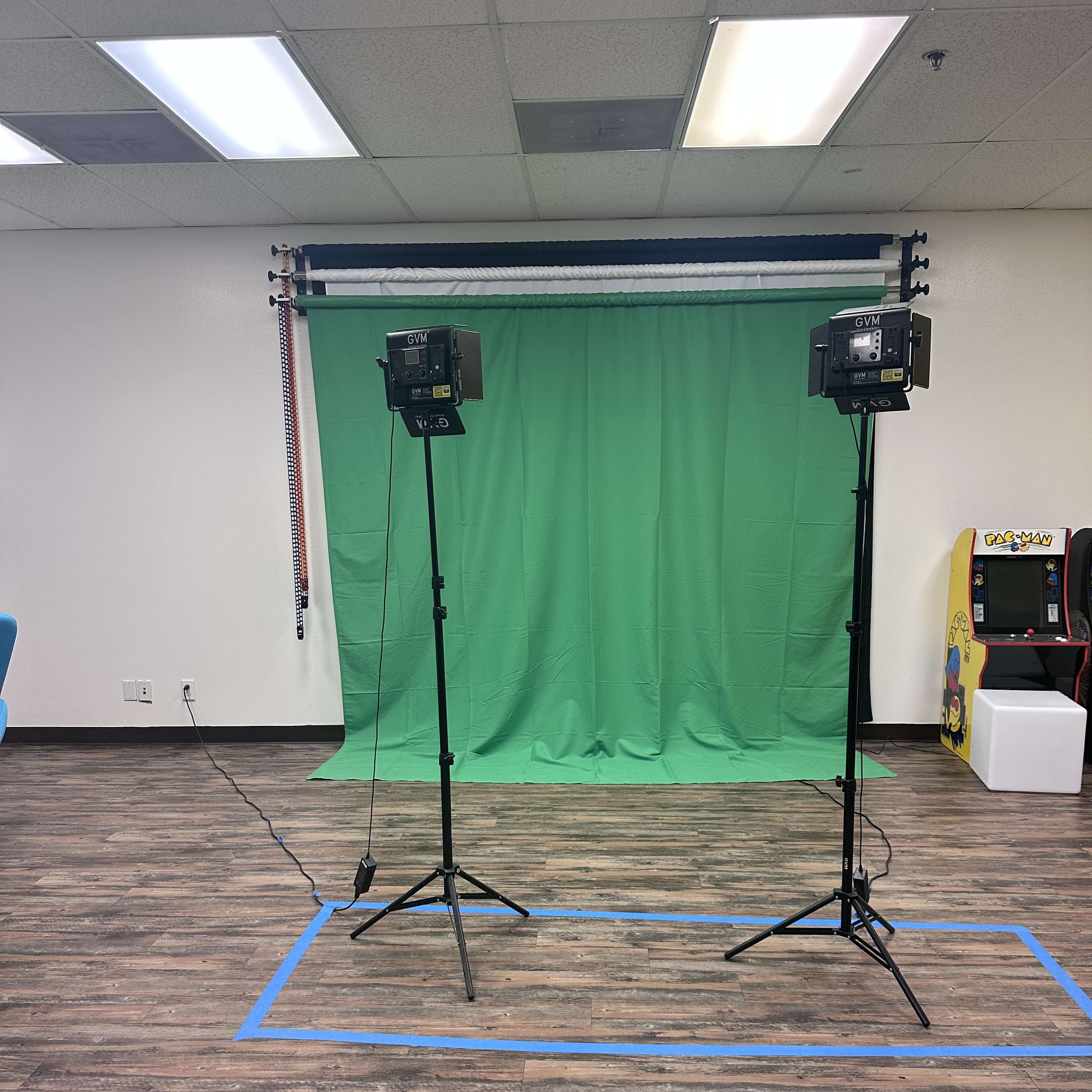 A room with a green screen setup flanked by two lighting stands, and an arcade machine in the background.