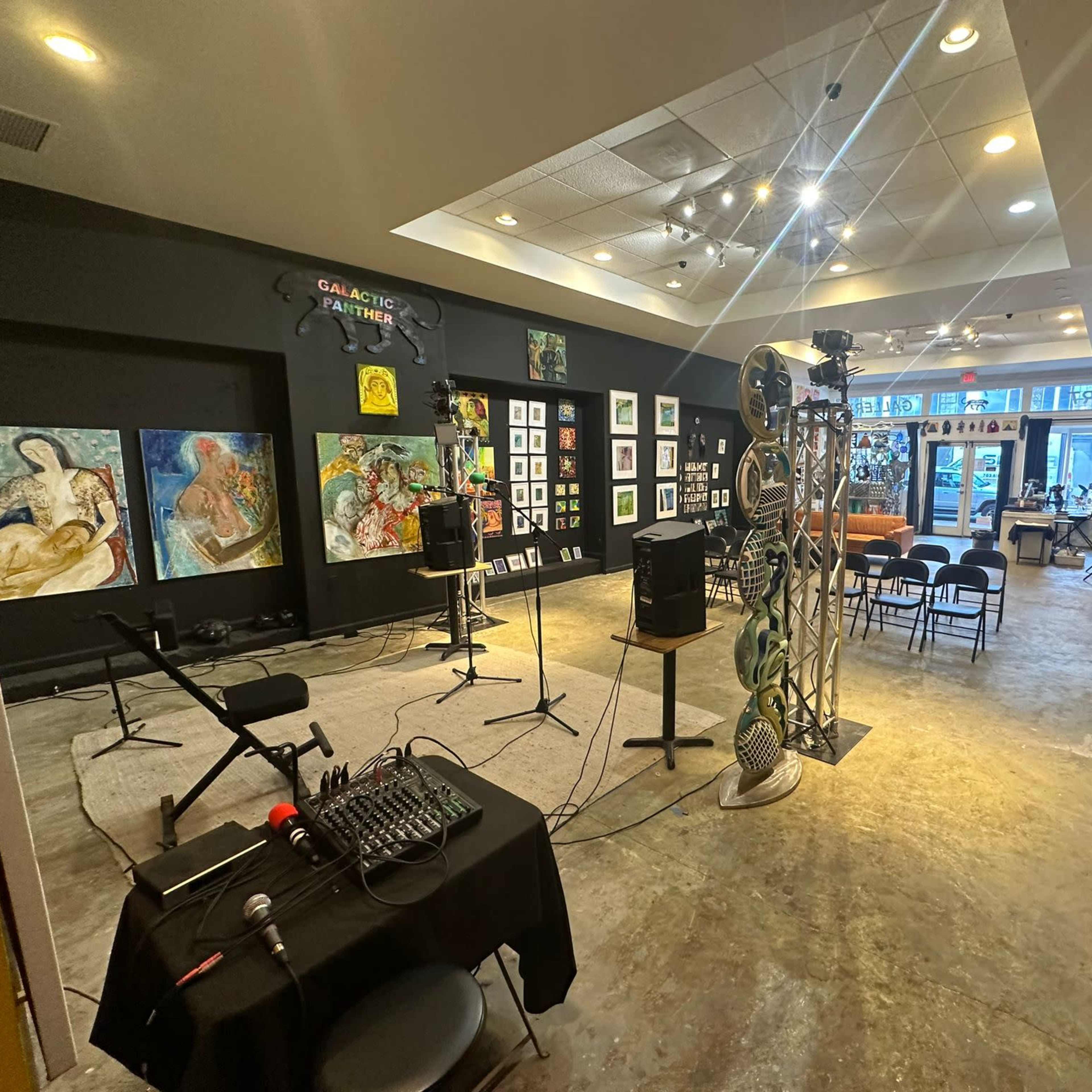 An art gallery and performance space with a stage setup, framed artworks on the walls, and chairs arranged for an audience.