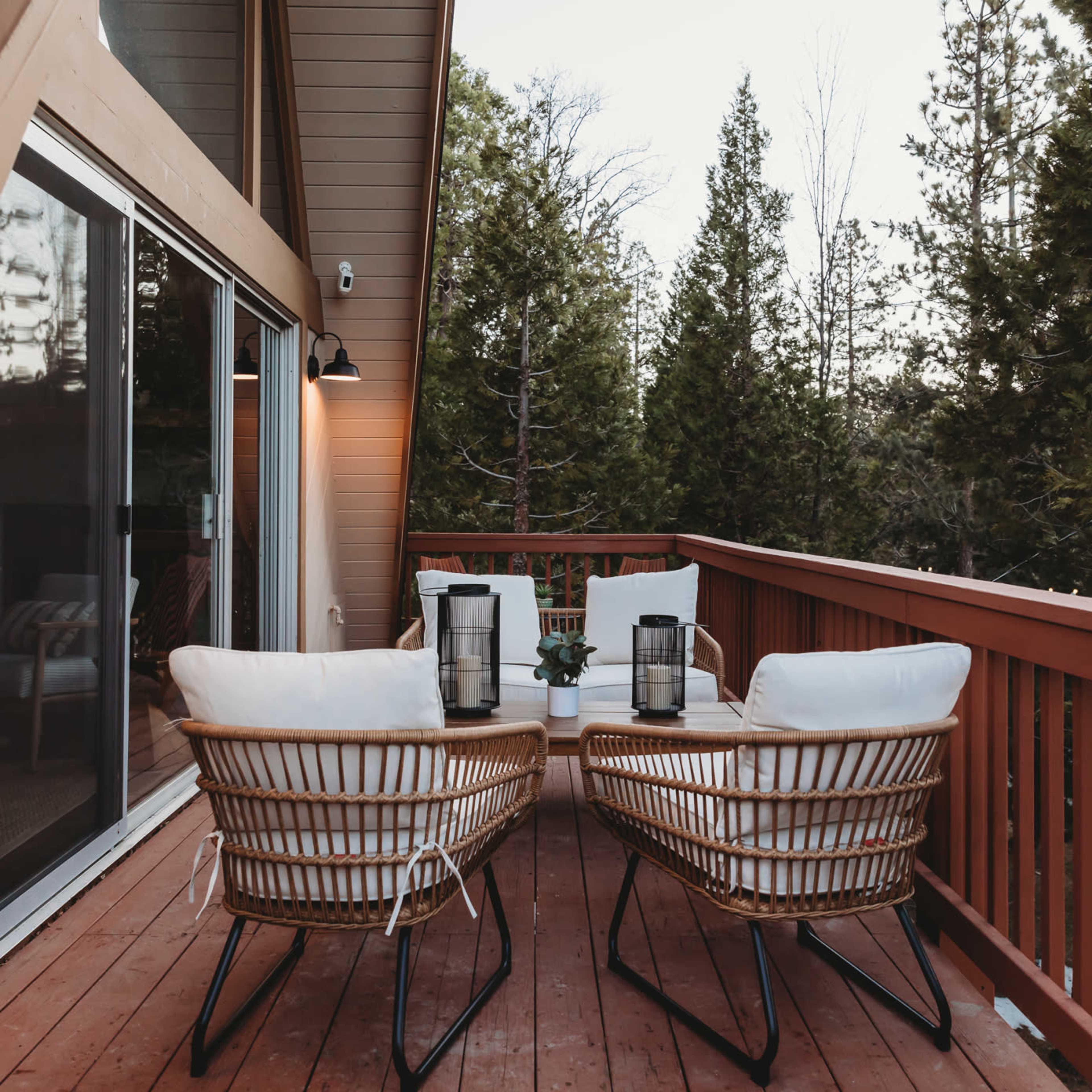 A wooden deck features two rattan chairs and a table, surrounded by tall trees and a glass sliding door.
