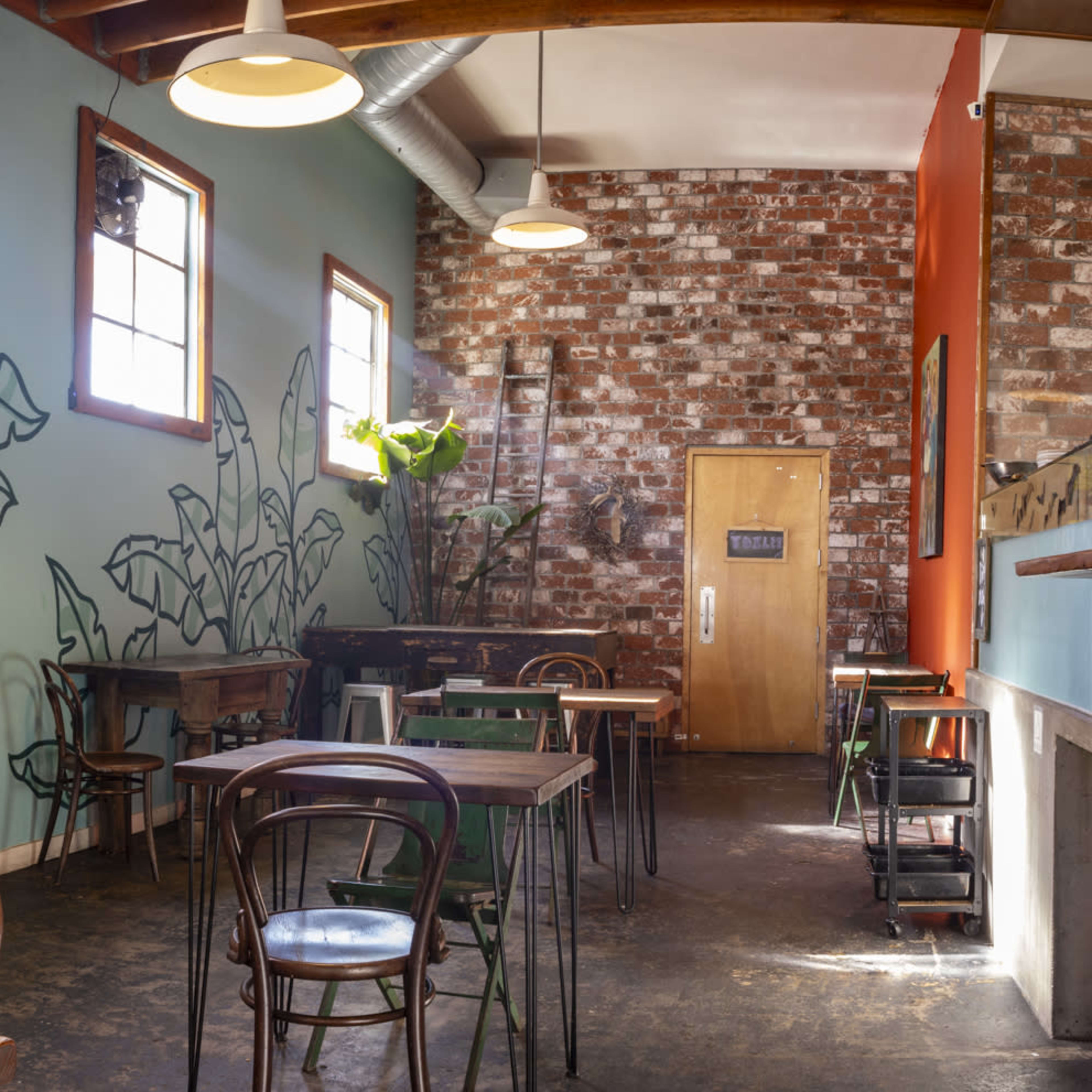 The interior of a cafe features wooden tables, a brick wall, and murals of large green leaves on the blue walls.