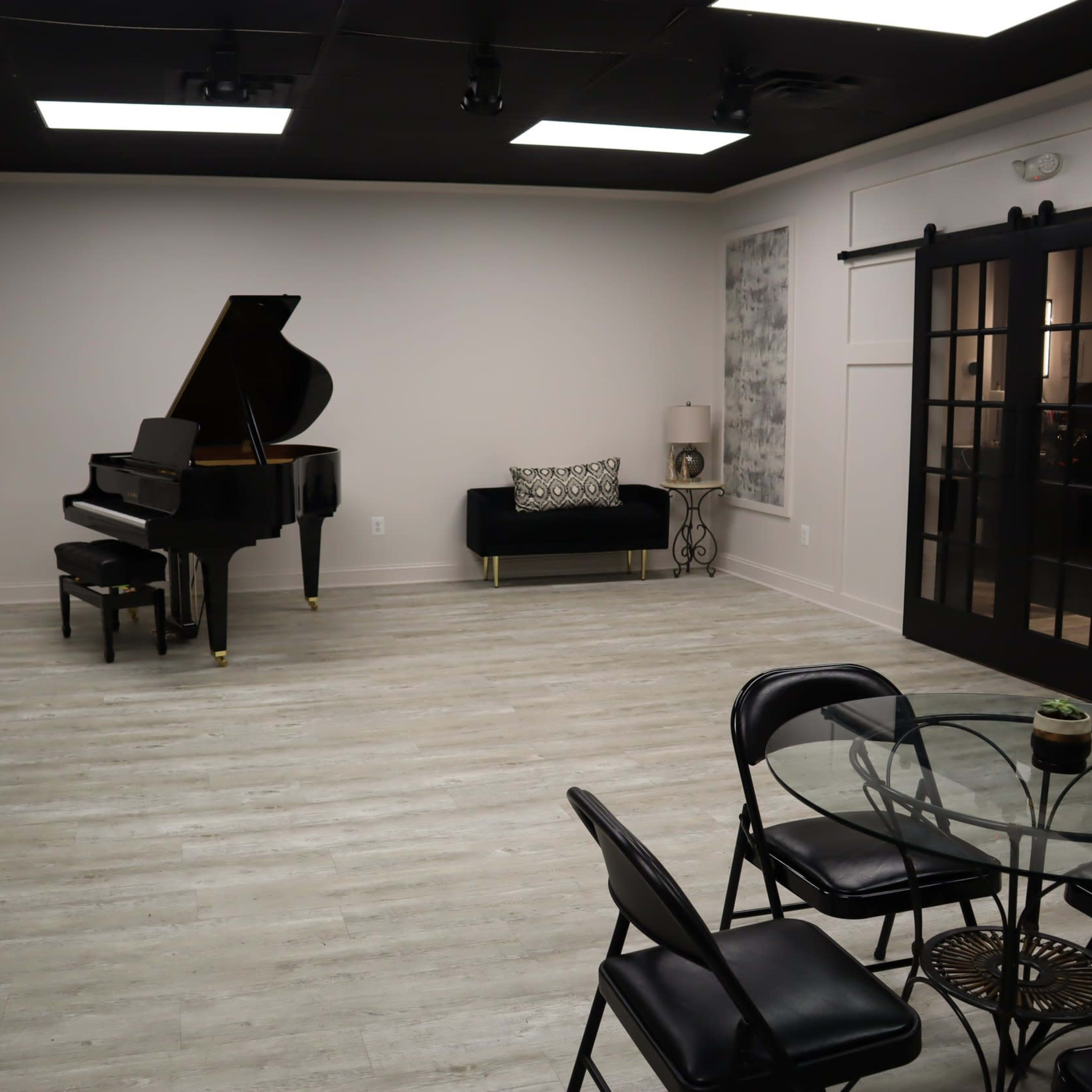 The room features a grand piano, a black sofa, a glass dining table with chairs, and large windows with black-framed doors.