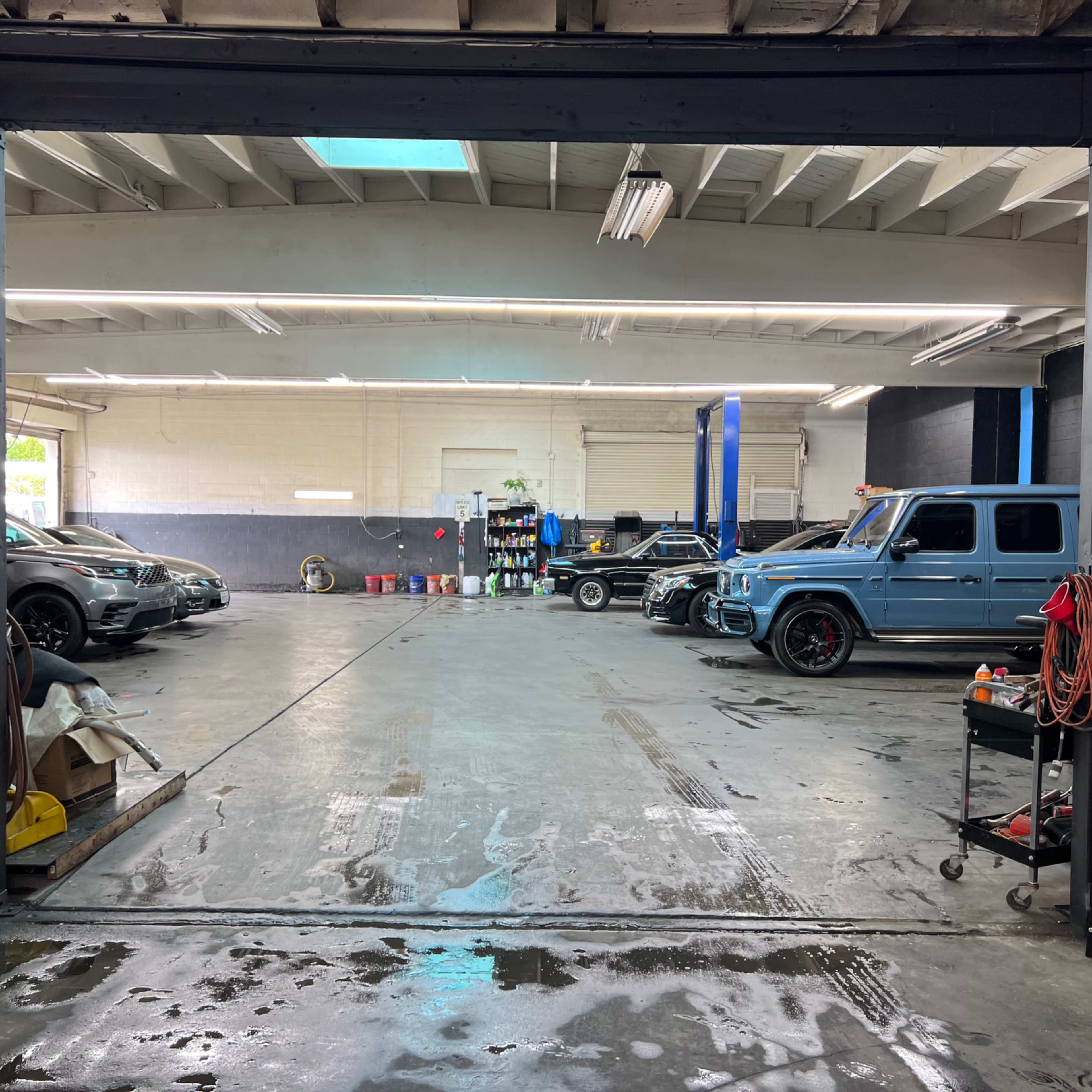 An automotive workshop with several vehicles parked inside, tools and supplies organized along the walls, and a lift in the background.