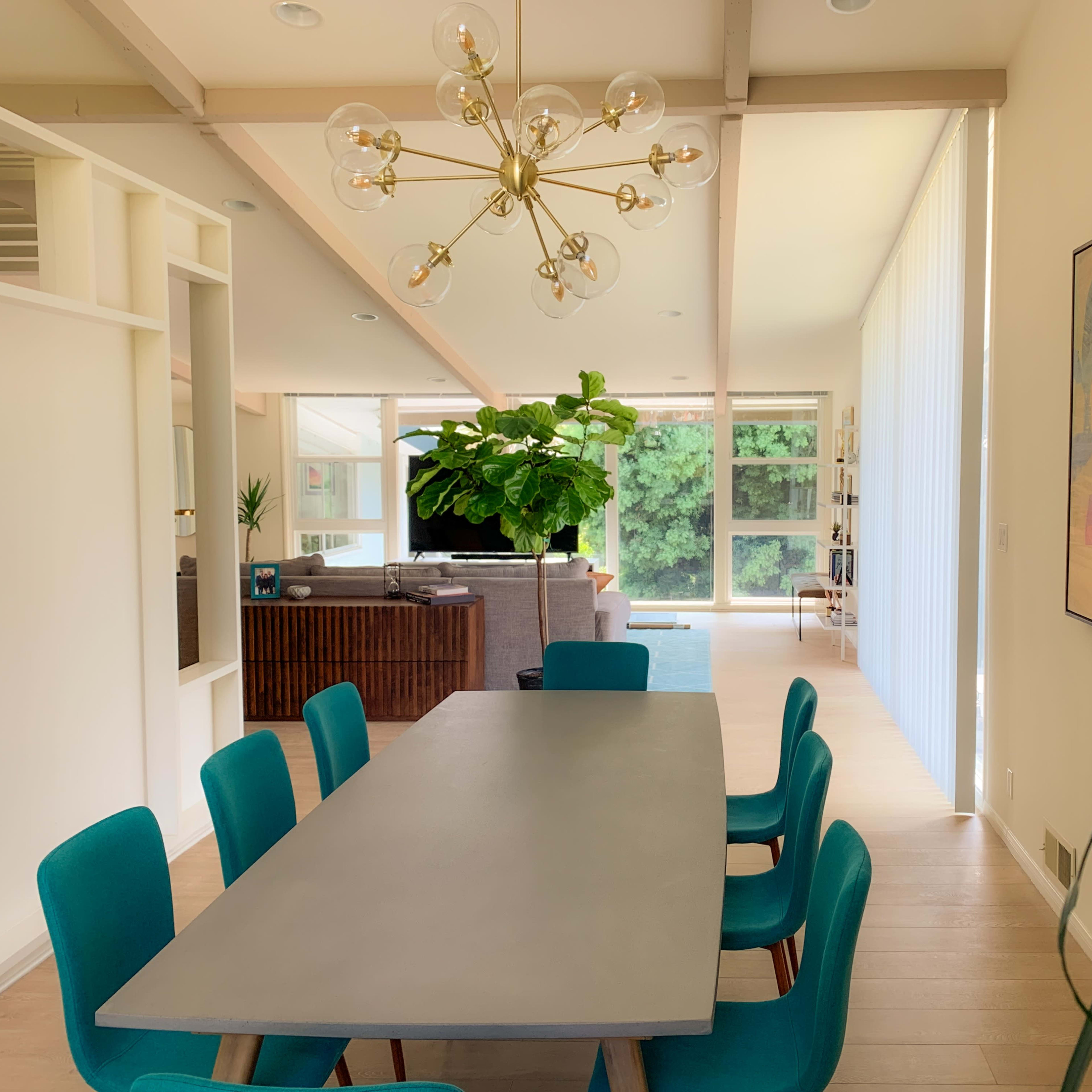 A modern dining area with a long gray table surrounded by teal chairs, leading into a living space with large windows and a prominent indoor plant.
