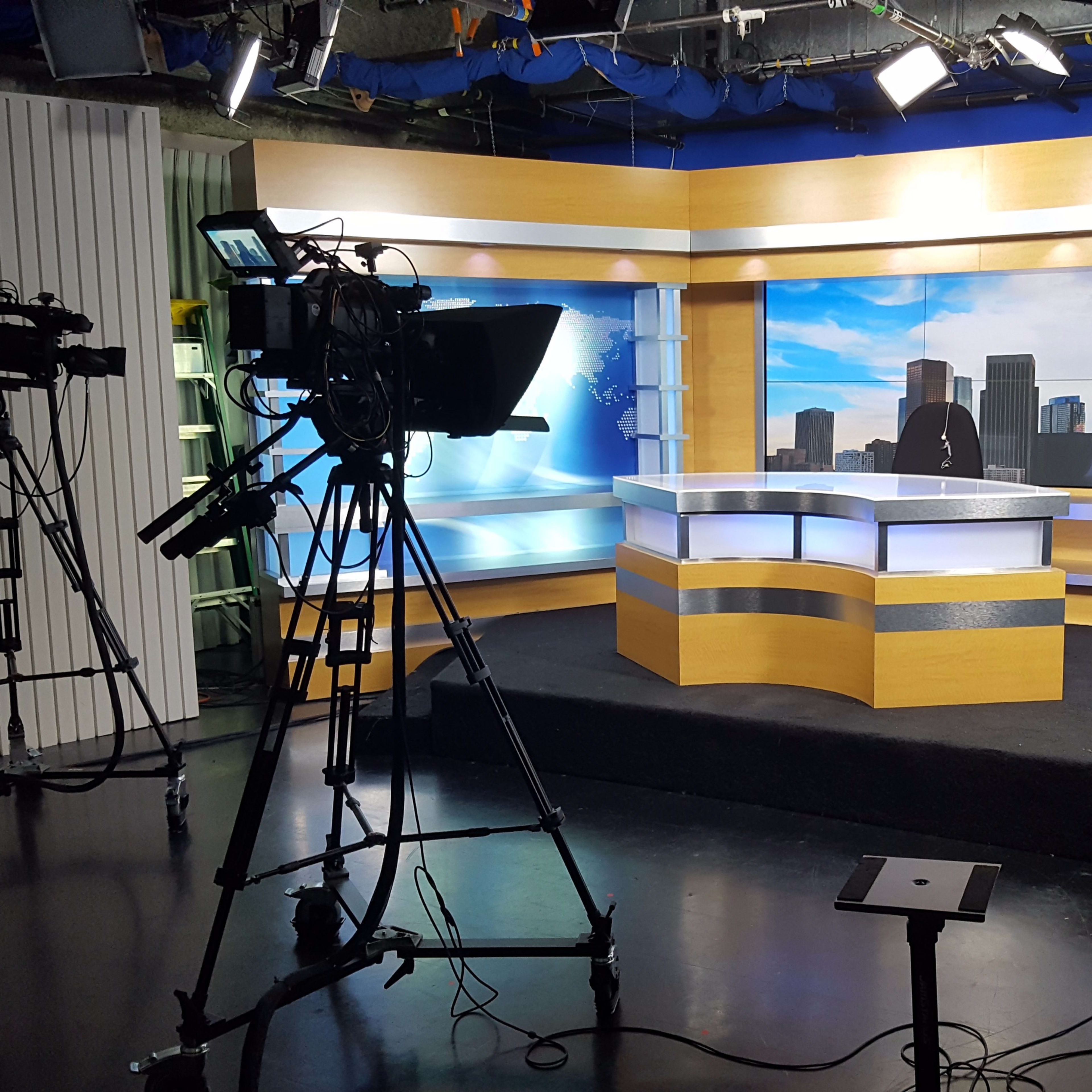 A television news studio with multiple cameras and a central news desk set against a city skyline backdrop.