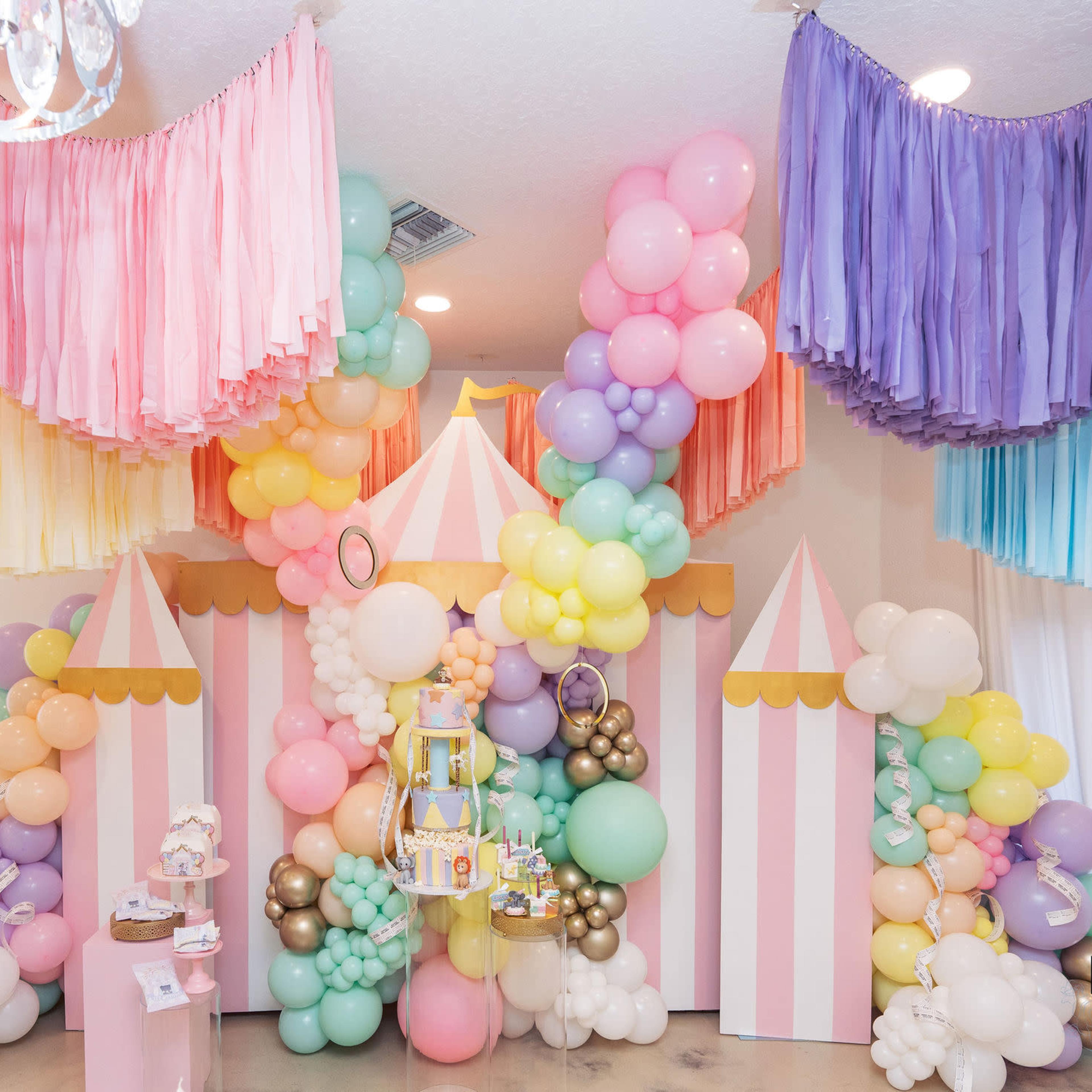 A festive party setup featuring a colorful balloon arch and a decorated backdrop resembling a circus or carnival theme, complete with pastel colors and striped elements.