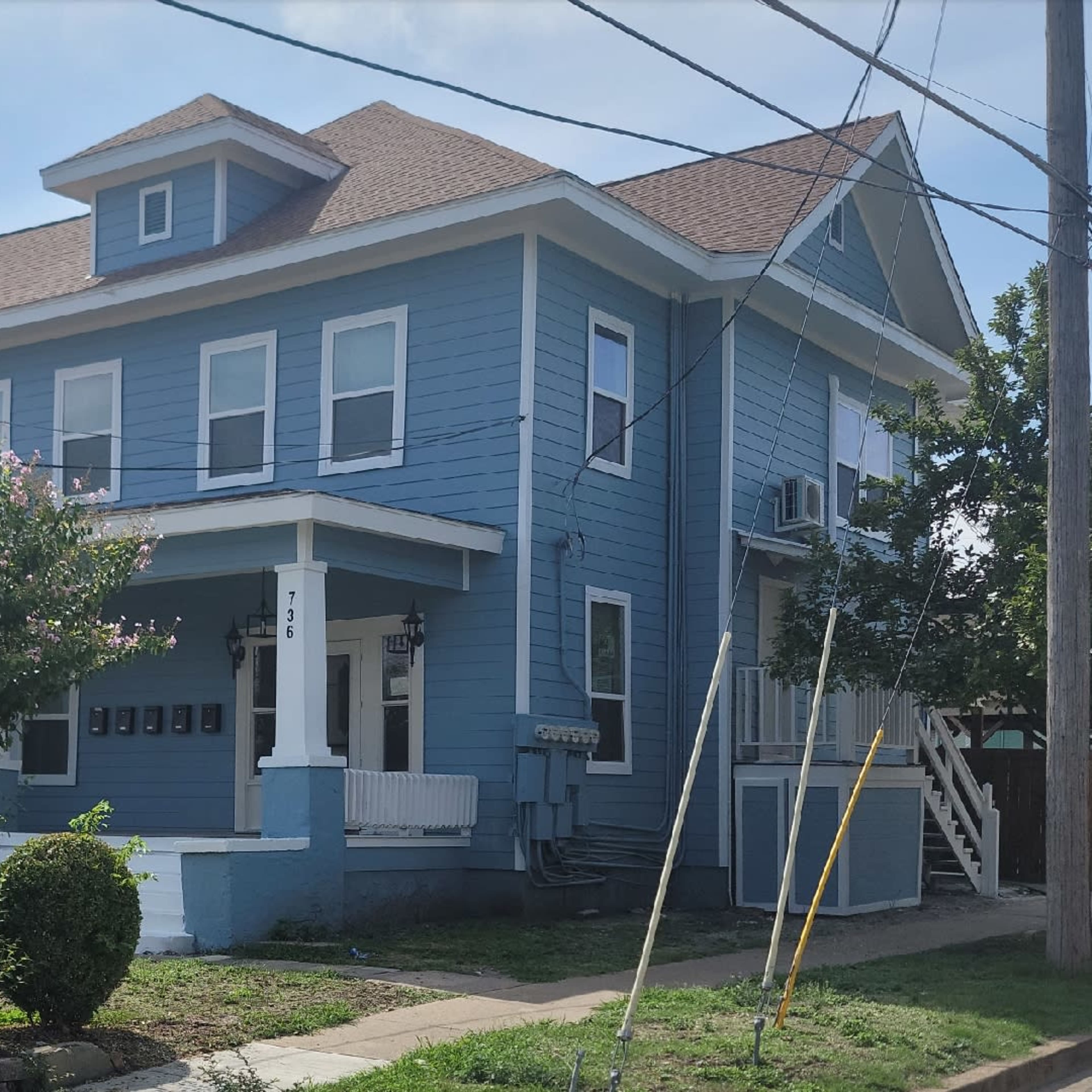 A two-story blue house with a porch and trees in front, situated on a residential street.