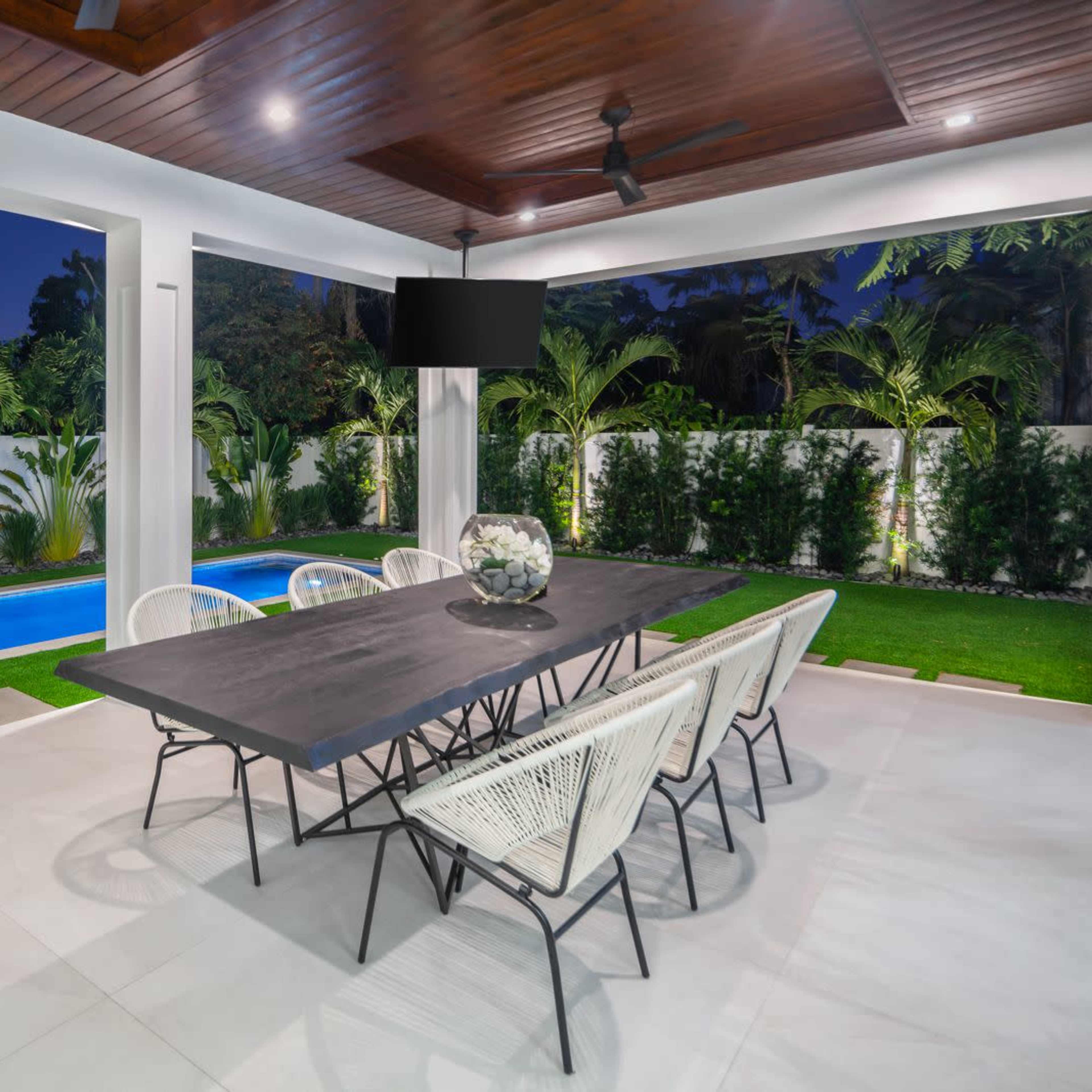 A sleek outdoor dining area features a large black table surrounded by white chairs, adjacent to a blue swimming pool and lush greenery, under a wooden ceiling with recessed lights.