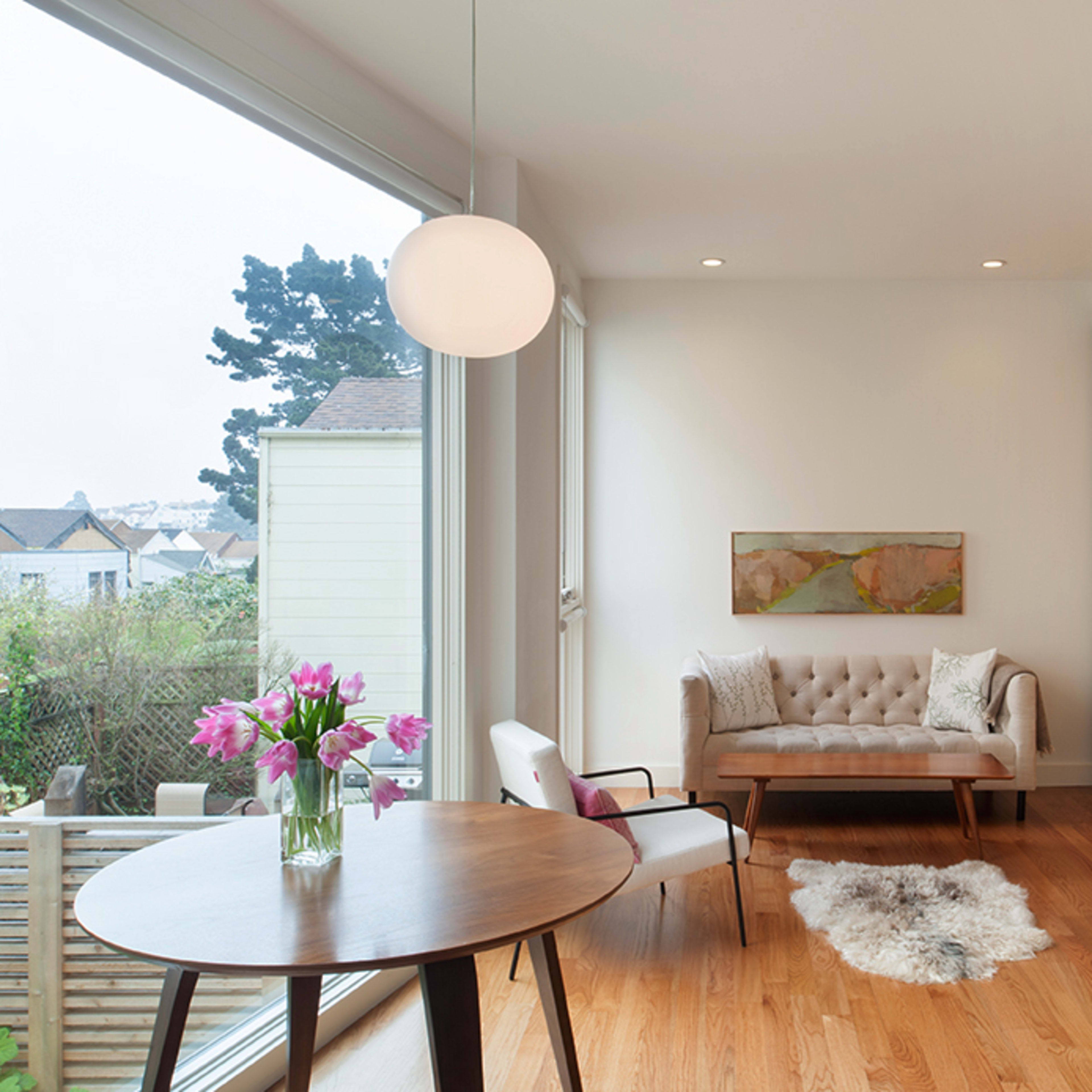 A cozy living space features a light-colored sofa, a round table with a vase of flowers, and large windows allowing natural light to enter.