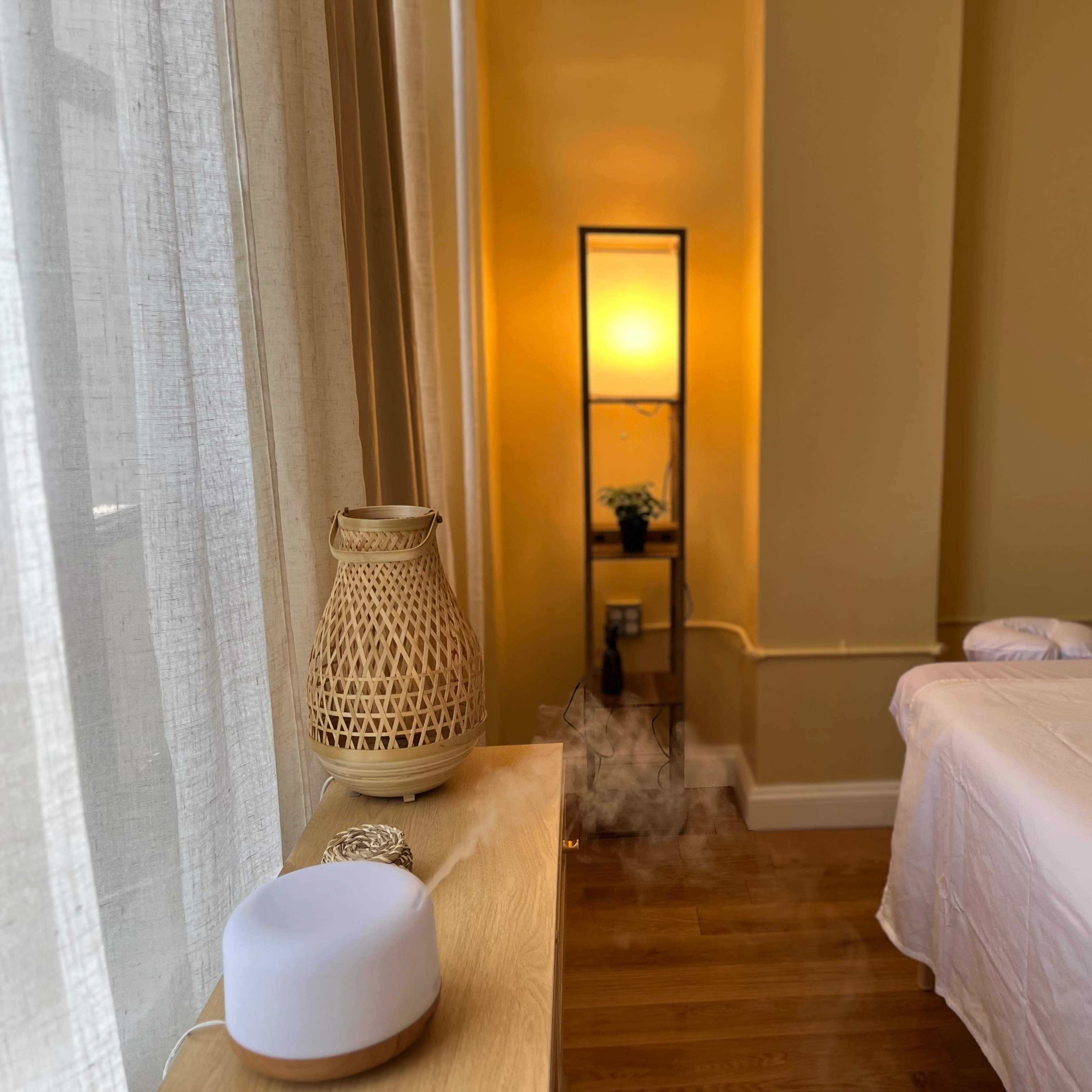 A calm spa room featuring a massage table, a decorative lamp, and a diffuser emitting mist near a window draped with sheer curtains.
