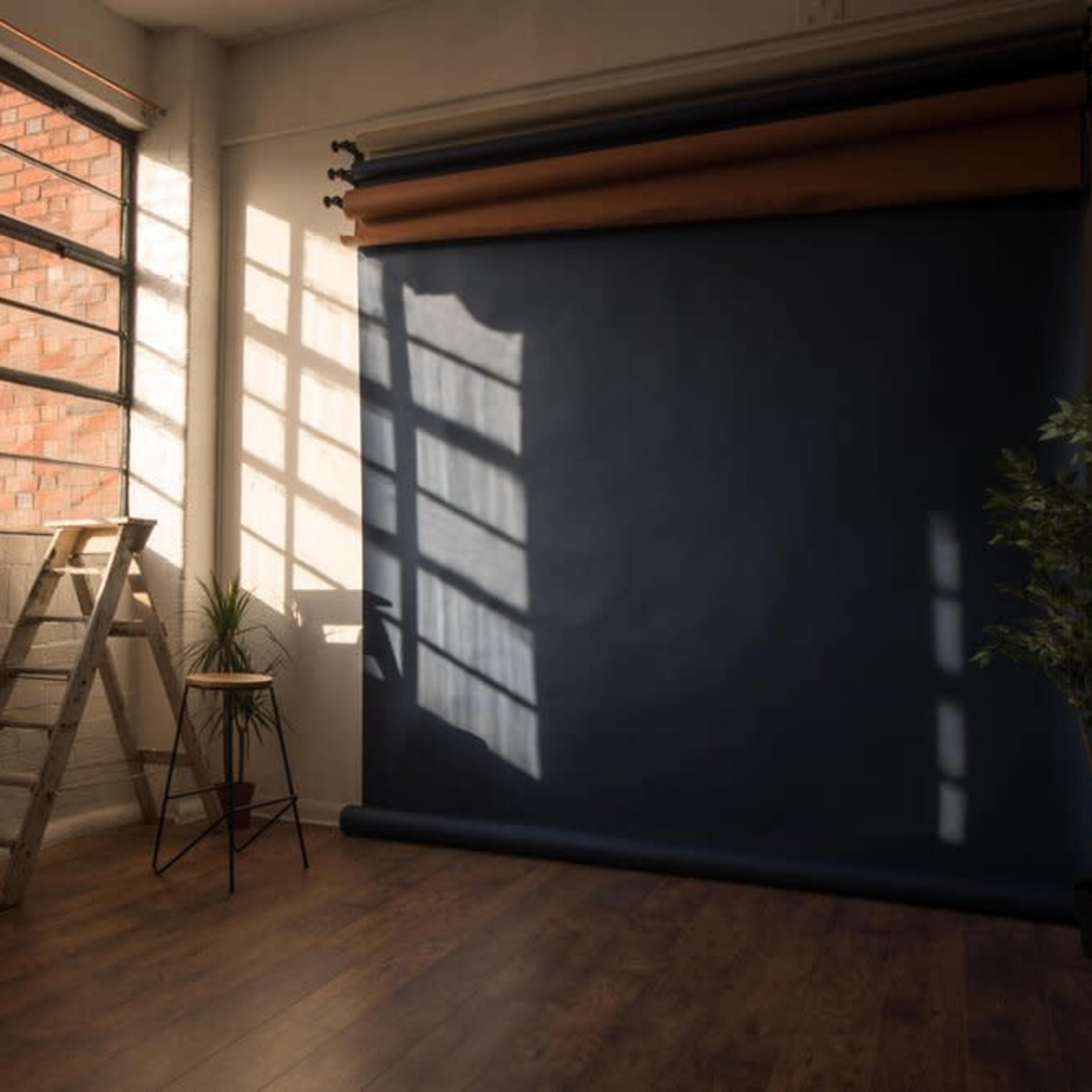 A spacious interior with a large window, a wooden ladder, a stool, a potted plant, and a dark backdrop rolled down against a wall.