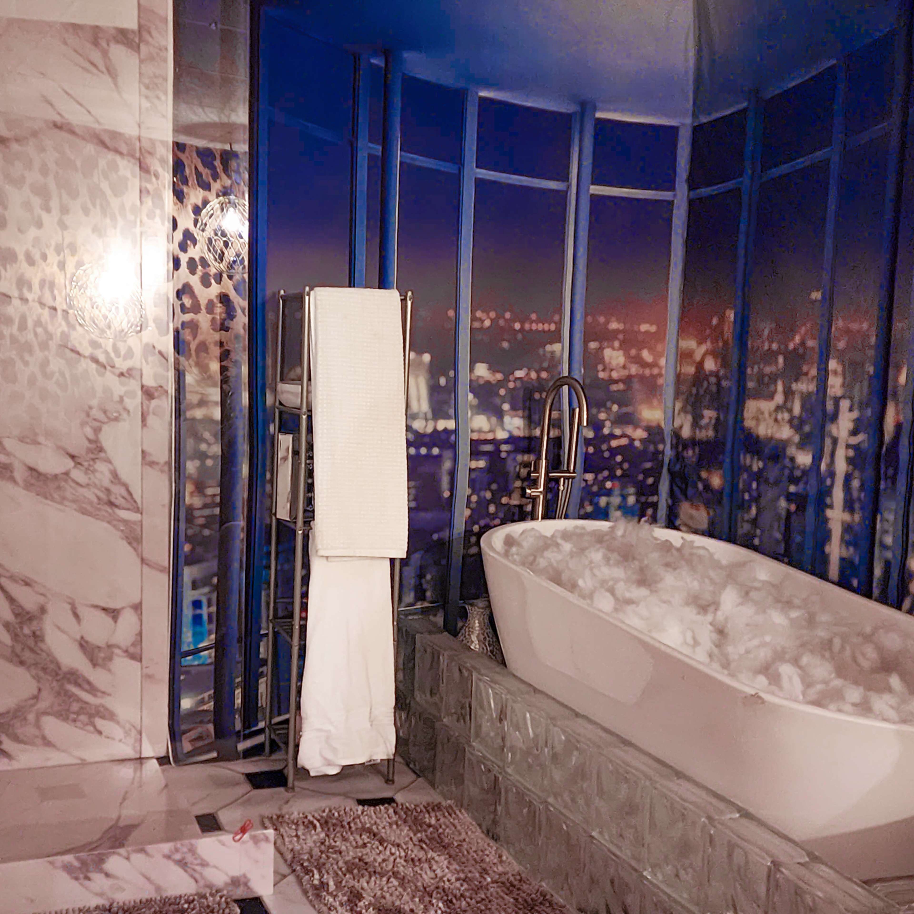 A modern bathroom with a freestanding bathtub surrounded by stone walls, a glass block base, and a backdrop of a city skyline at night.