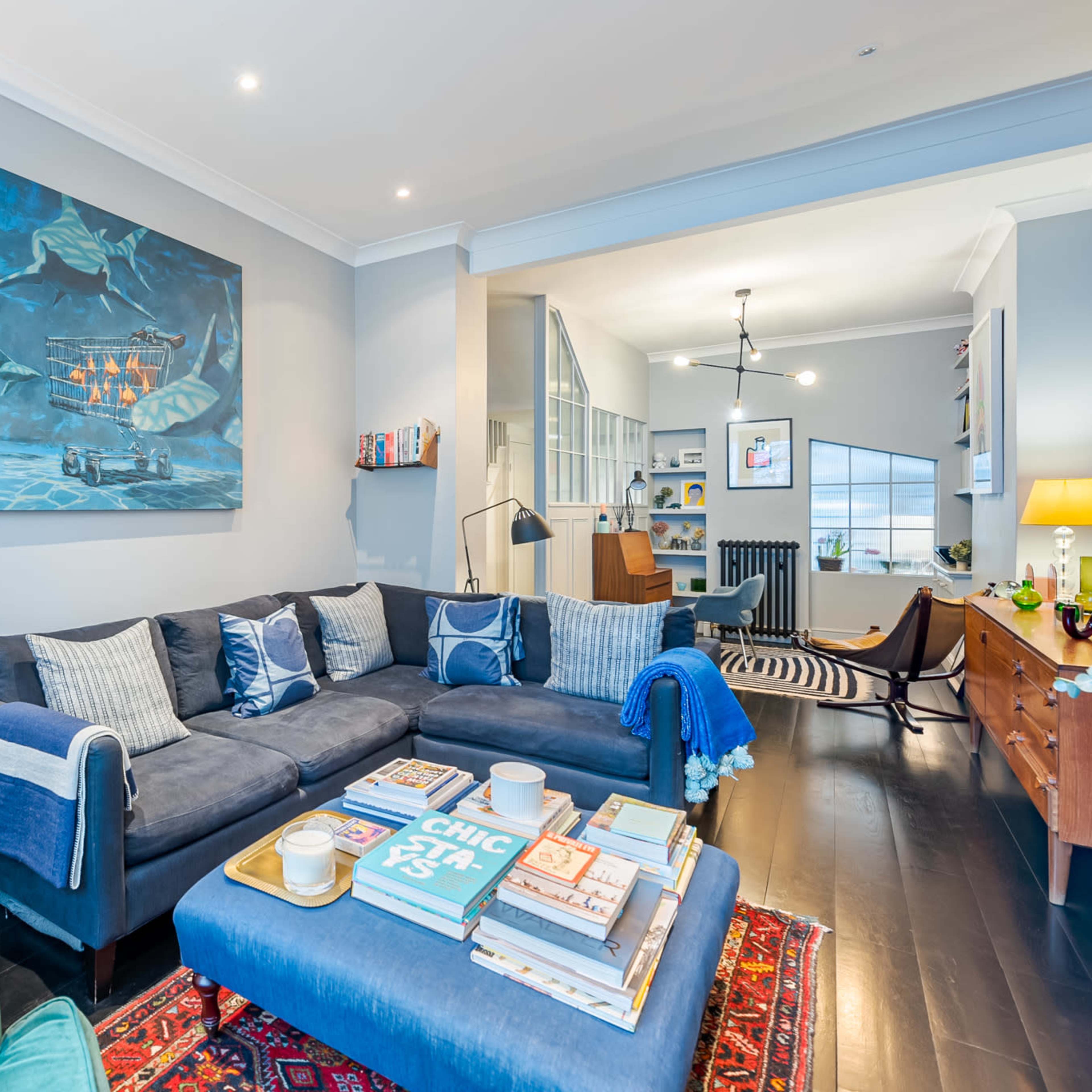 A modern living room features a dark blue sectional sofa with patterned cushions, a large artwork on the wall, and a coffee table stacked with books and decor.