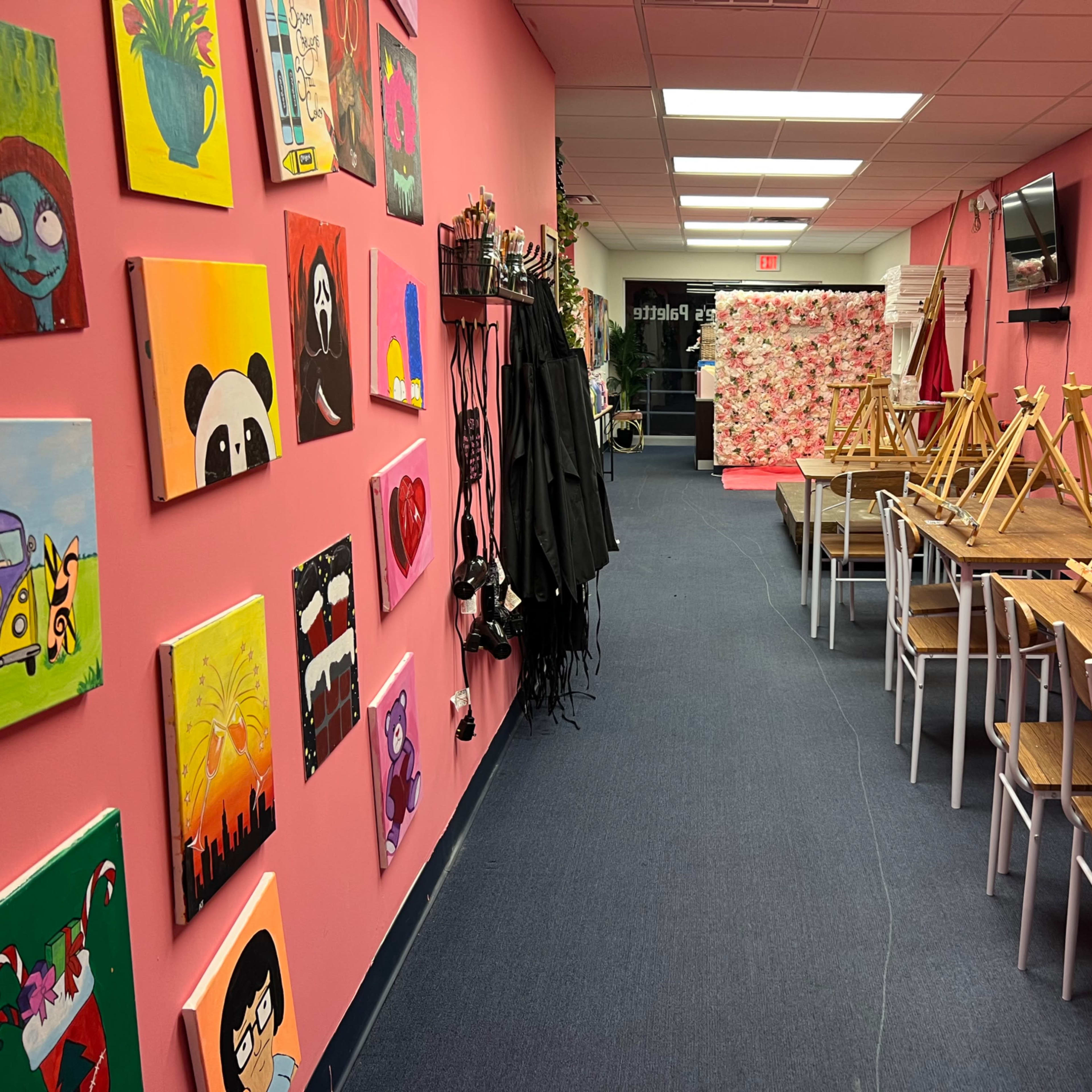 An art studio with pink walls, decorated with various paintings, featuring empty tables and easels arranged along the sides.