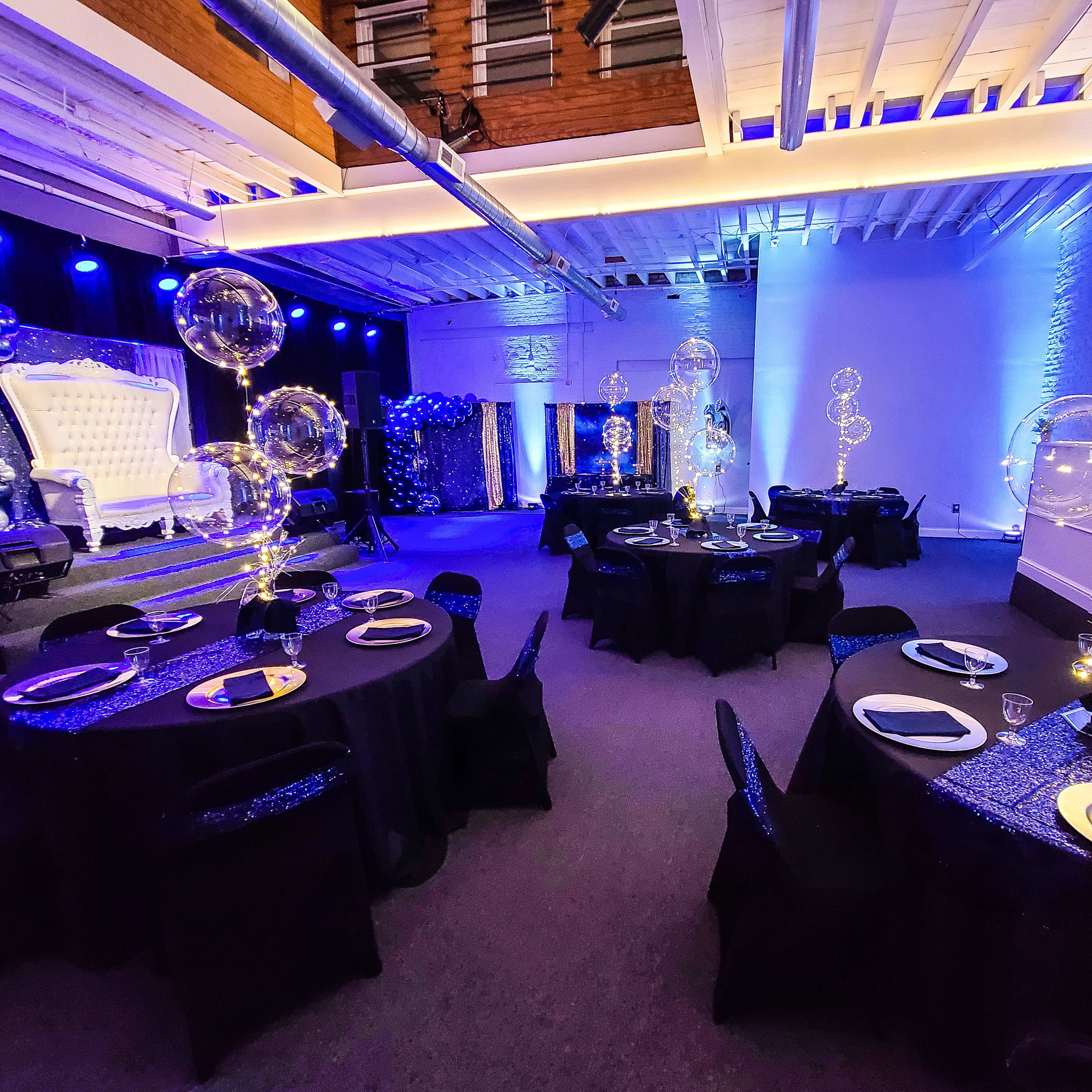 The venue features round tables with black tablecloths and elegant centerpieces, surrounded by balloons and decorative lights in a dimly lit setting.