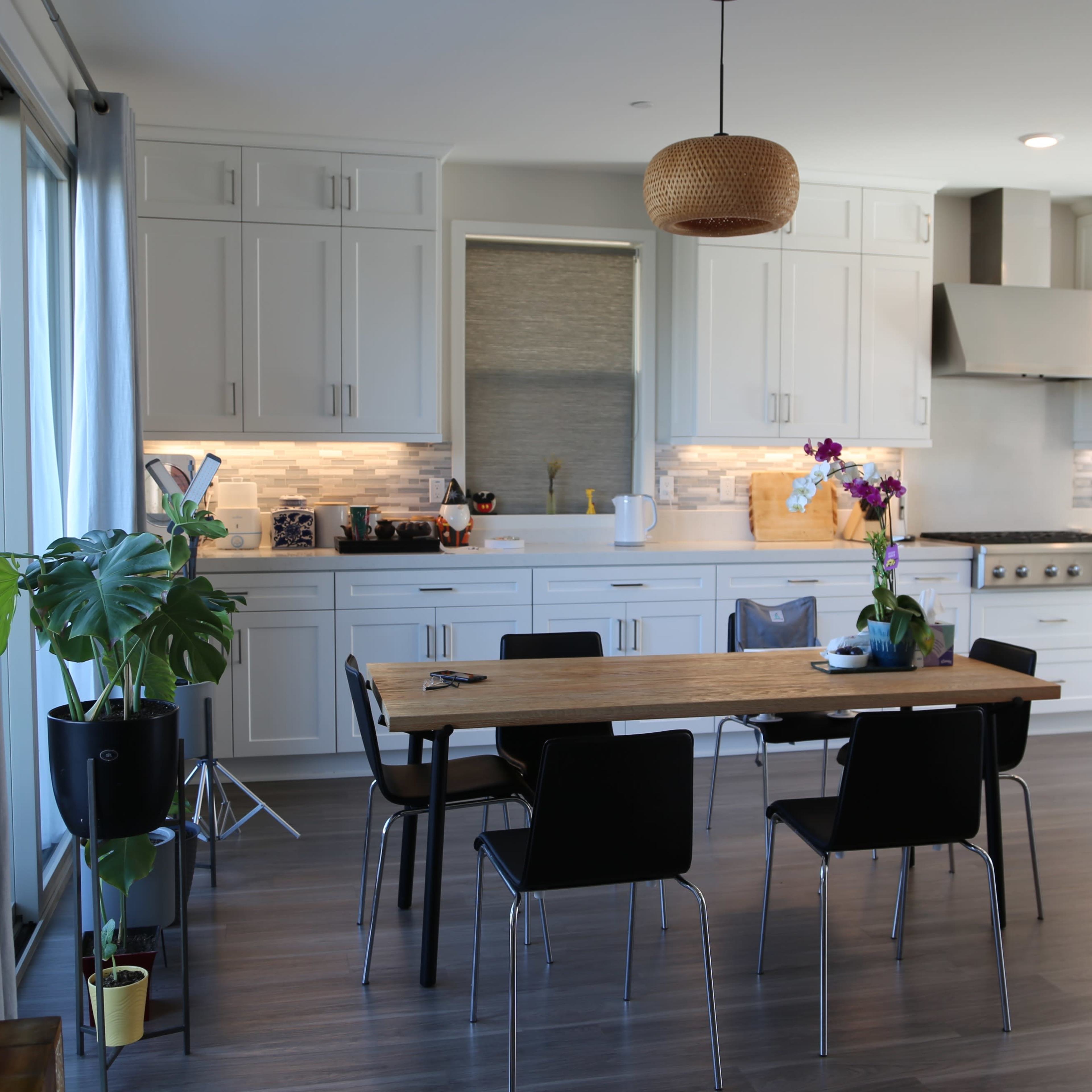 A modern kitchen features a long wooden dining table surrounded by black chairs, with white cabinetry and a large window in the background.