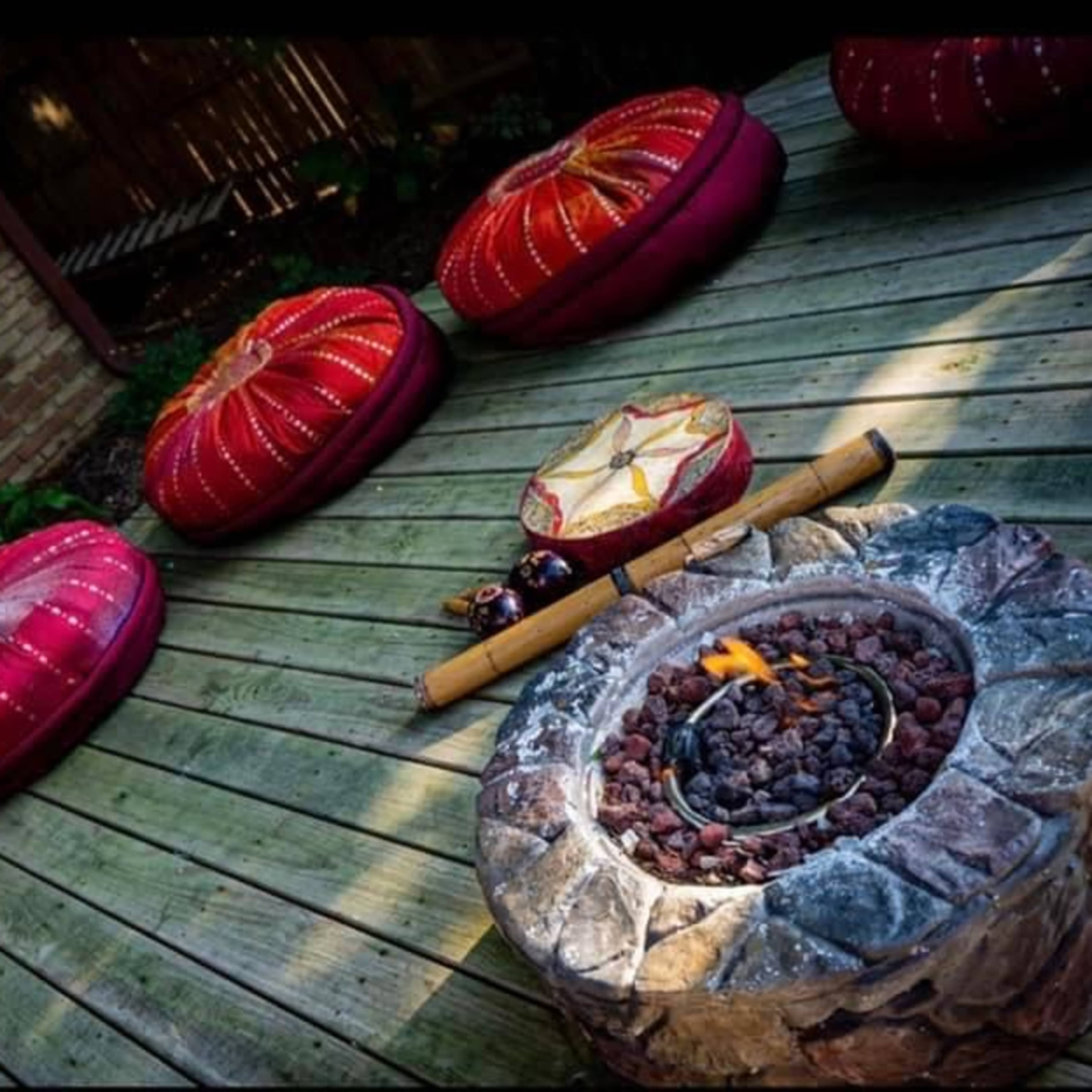A circular stone fire pit is surrounded by several round, red cushions on a wooden deck.