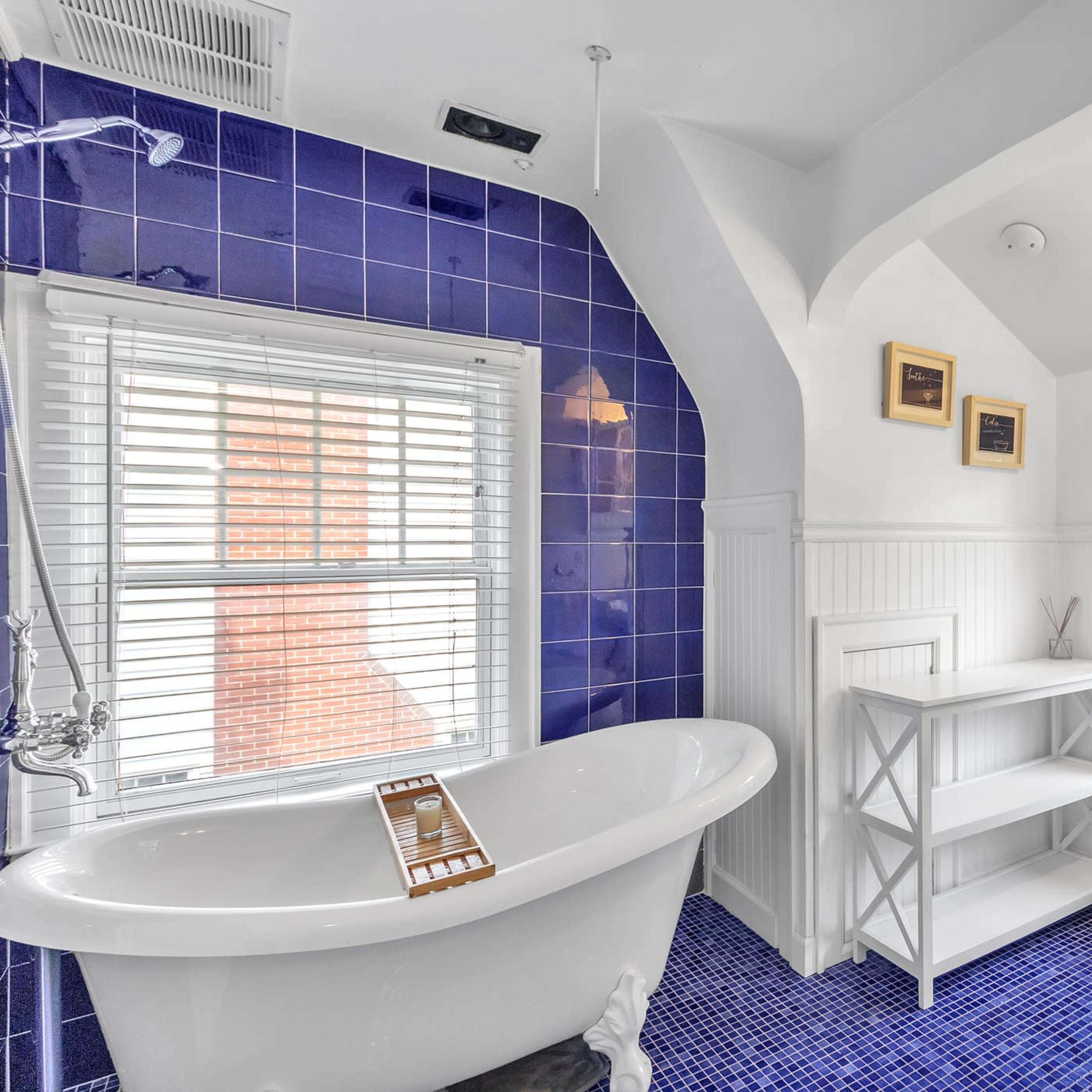 A bathroom with a freestanding bathtub, blue tile flooring, and a window overlooking a green outdoor area.