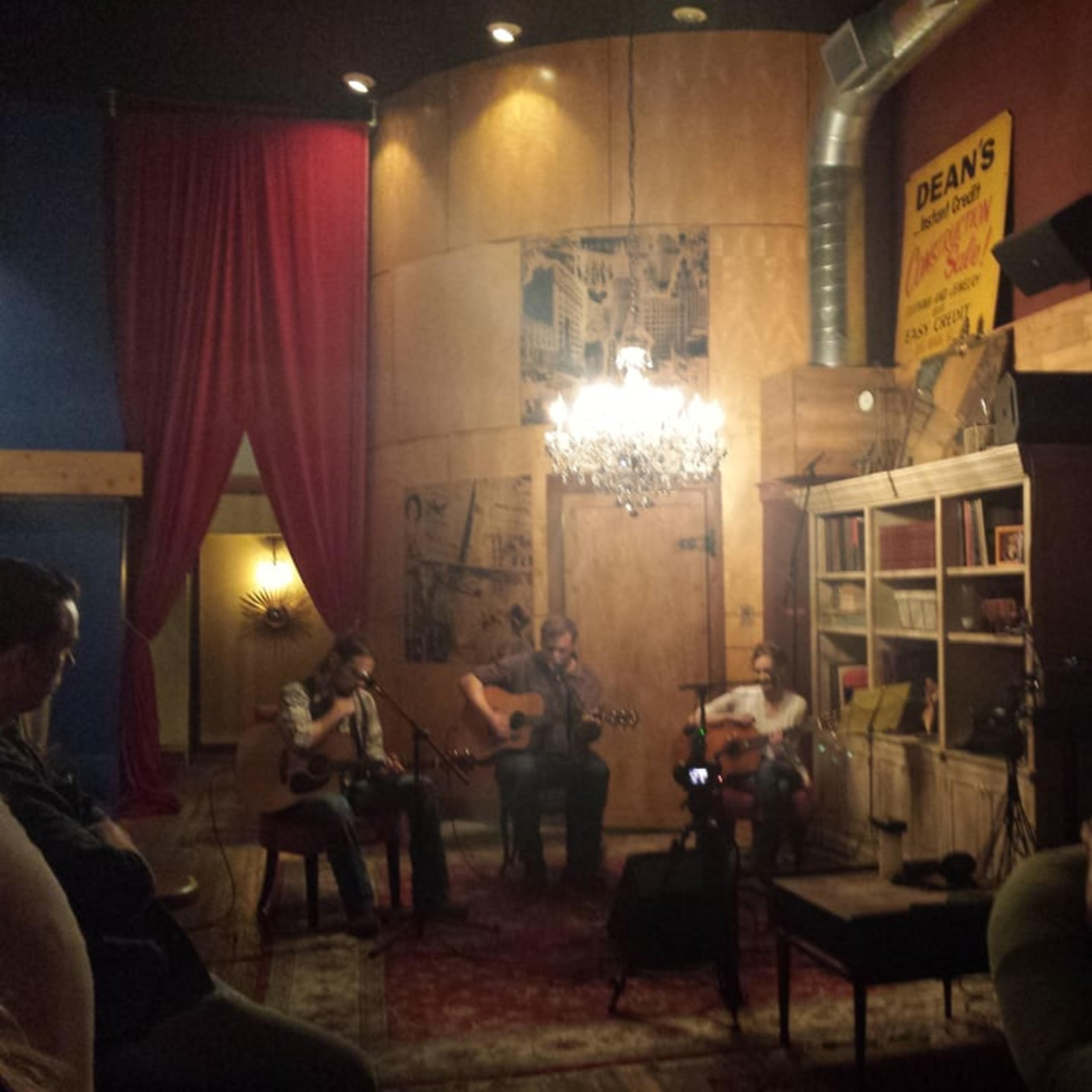A small crowd watches three musicians perform in an intimate venue with a chandelier and vintage decor.