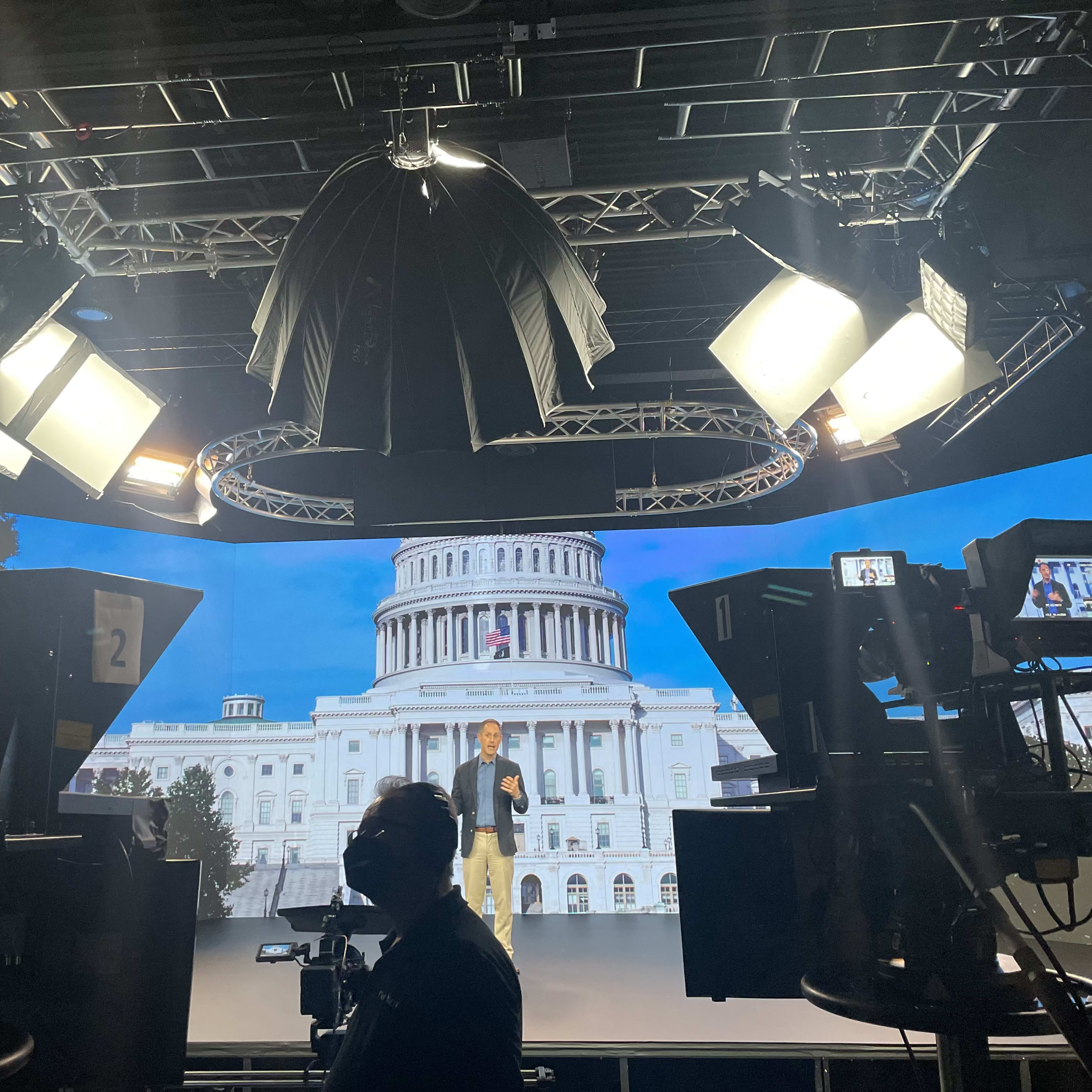 A man stands on a stage in a studio with the United States Capitol building projected in the background, surrounded by studio lights and cameras.
