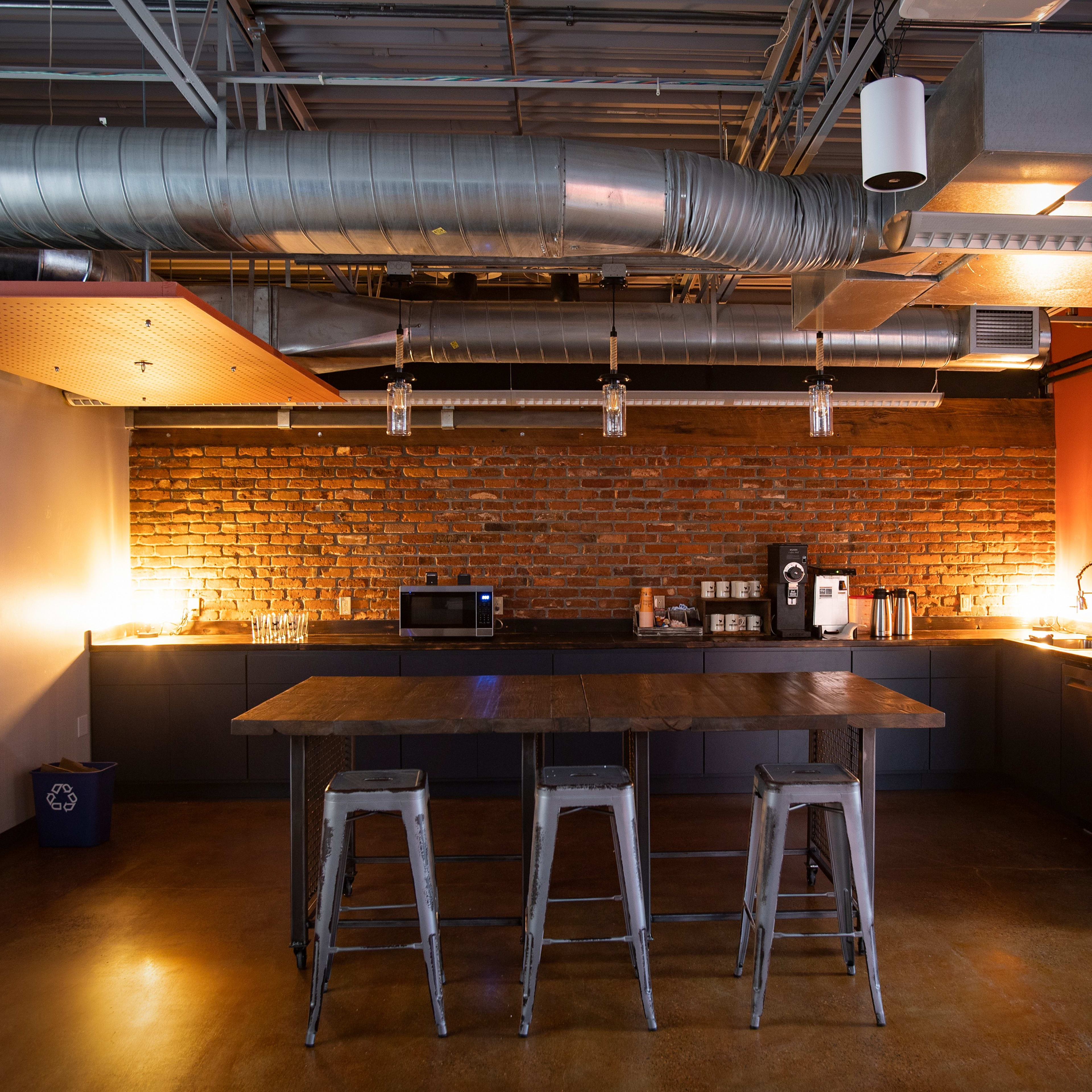 A modern kitchen area with a brick wall, stainless steel appliances, a large table, and metal stools.