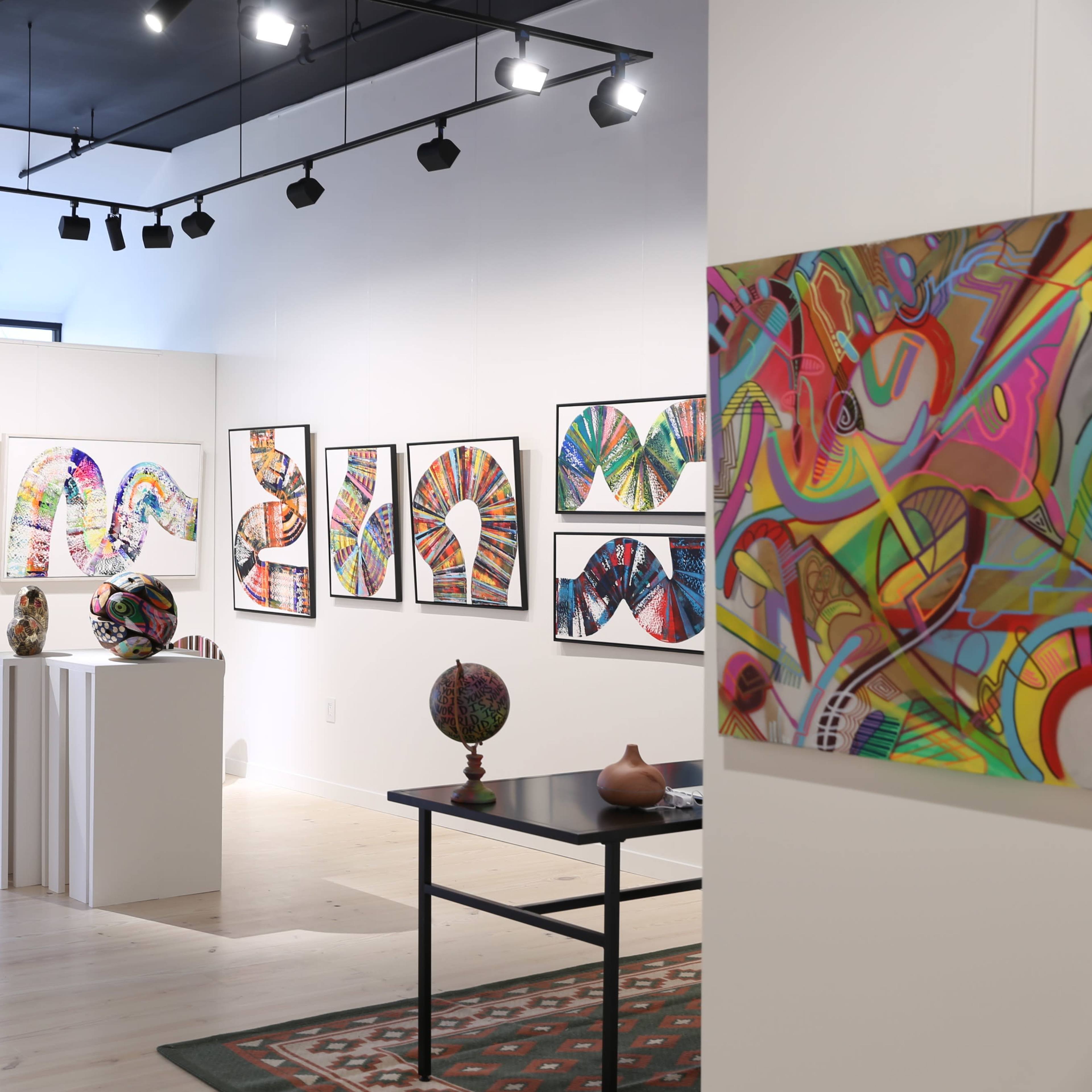 An art gallery interior featuring colorful abstract artworks displayed on the walls and various sculptures on pedestals and tables.