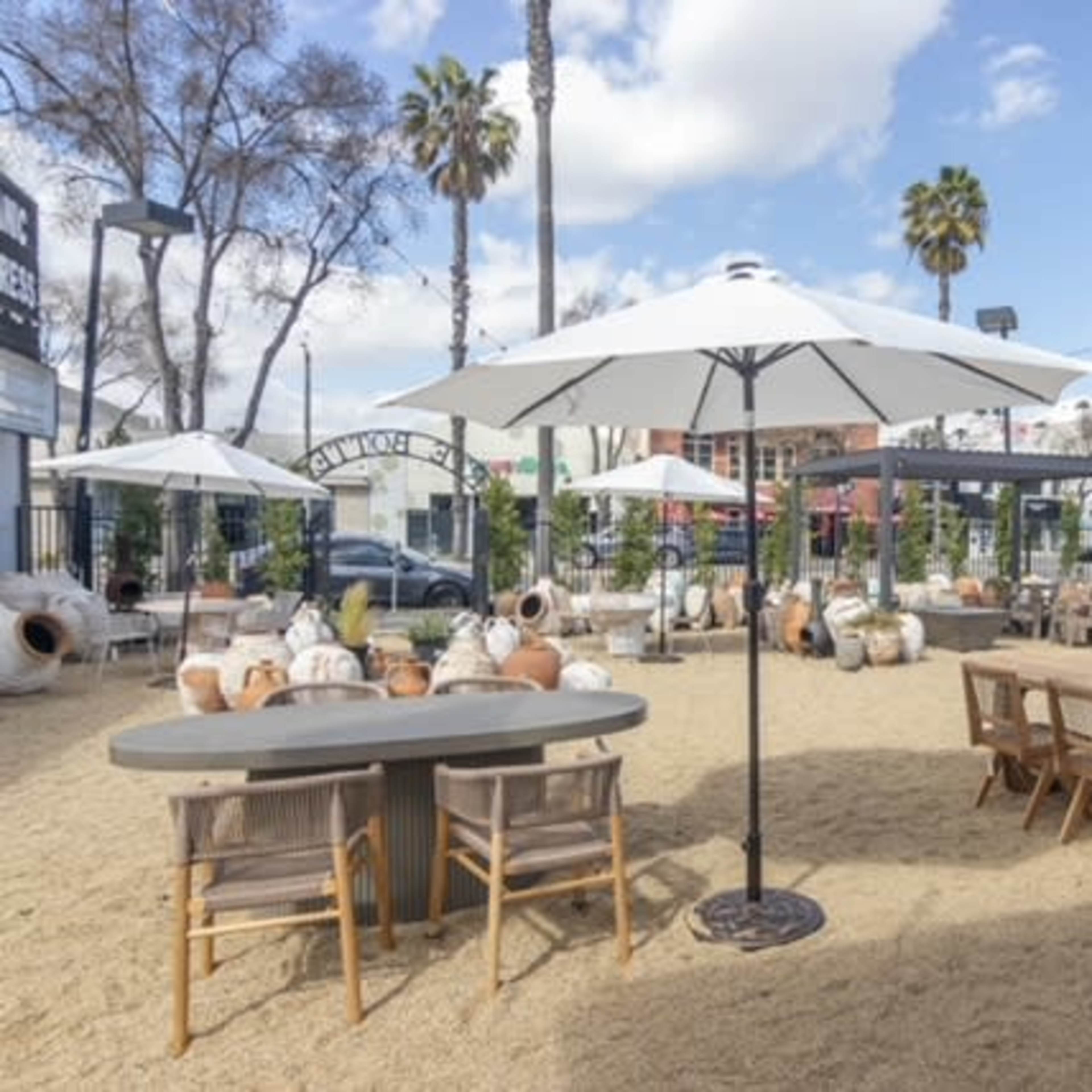 The scene features an outdoor dining area with wooden tables, chairs, and large patio umbrellas, surrounded by decorative pottery and palm trees under a clear sky.