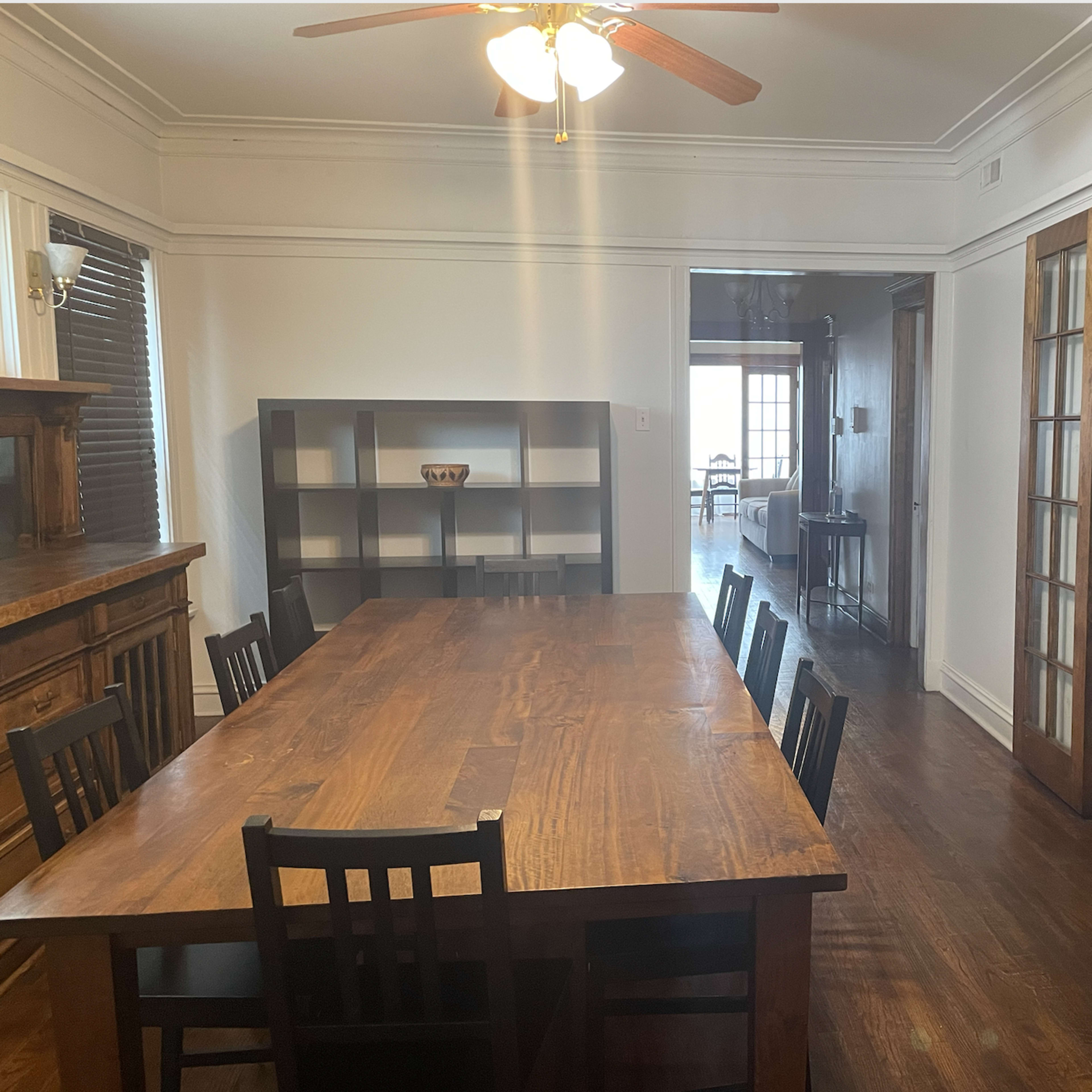 A large wooden dining table with black chairs is situated in a well-lit room, adjacent to an open doorway leading to another part of the house.