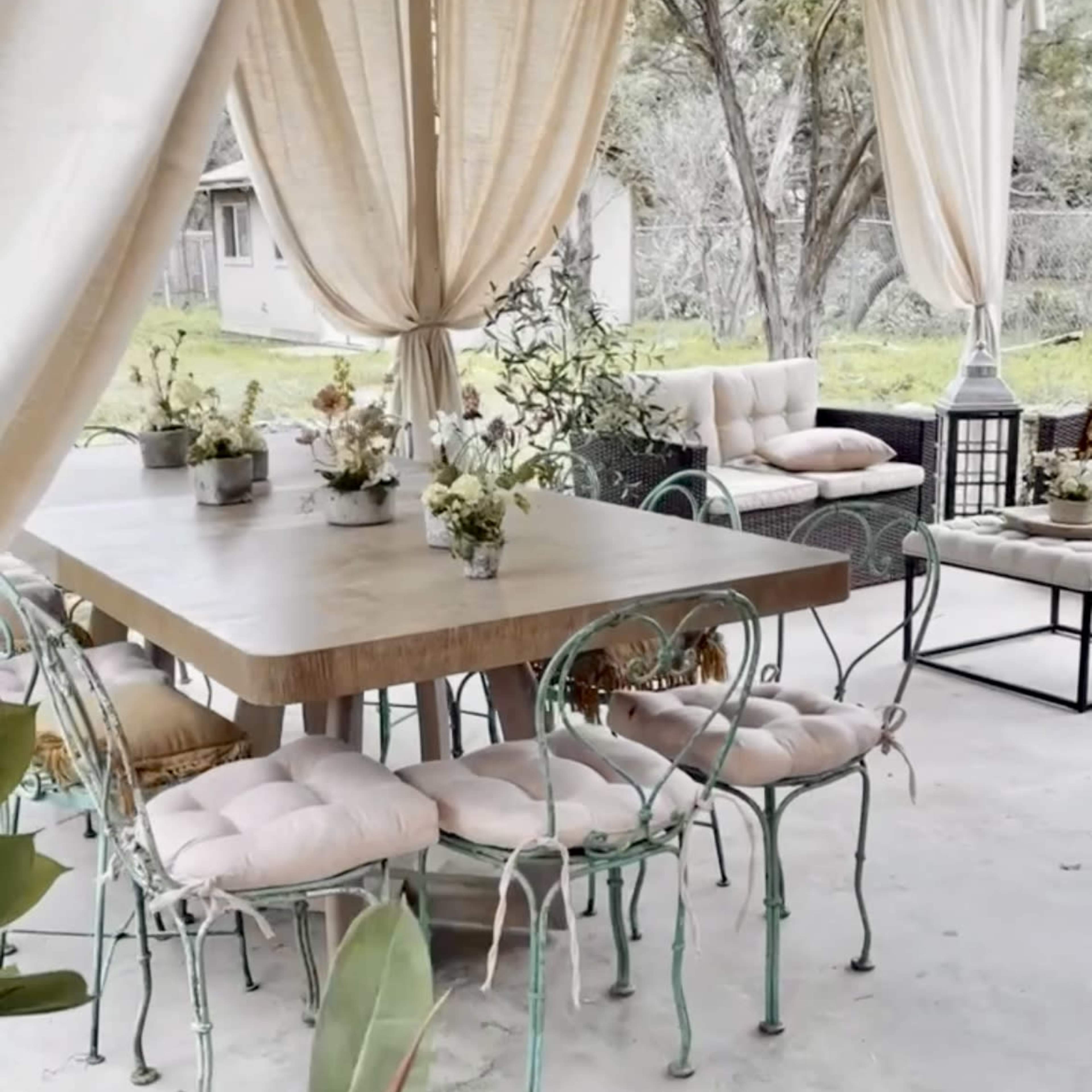 A covered outdoor seating area with a large wooden table surrounded by green metal chairs and potted plants.